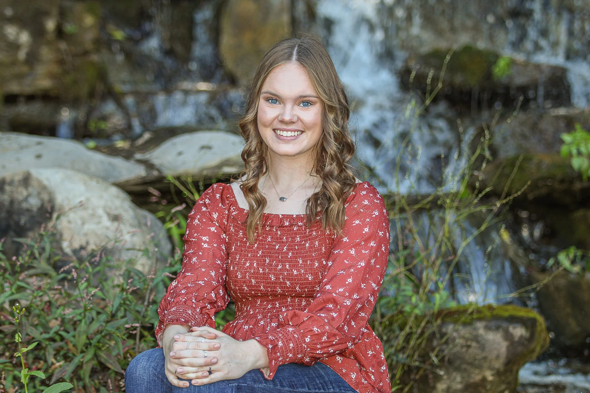 A smiling woman with blue eyes and long curly brown hair sitting outdoors by a stream in a natural setting, wearing a red floral blouse and blue jeans.