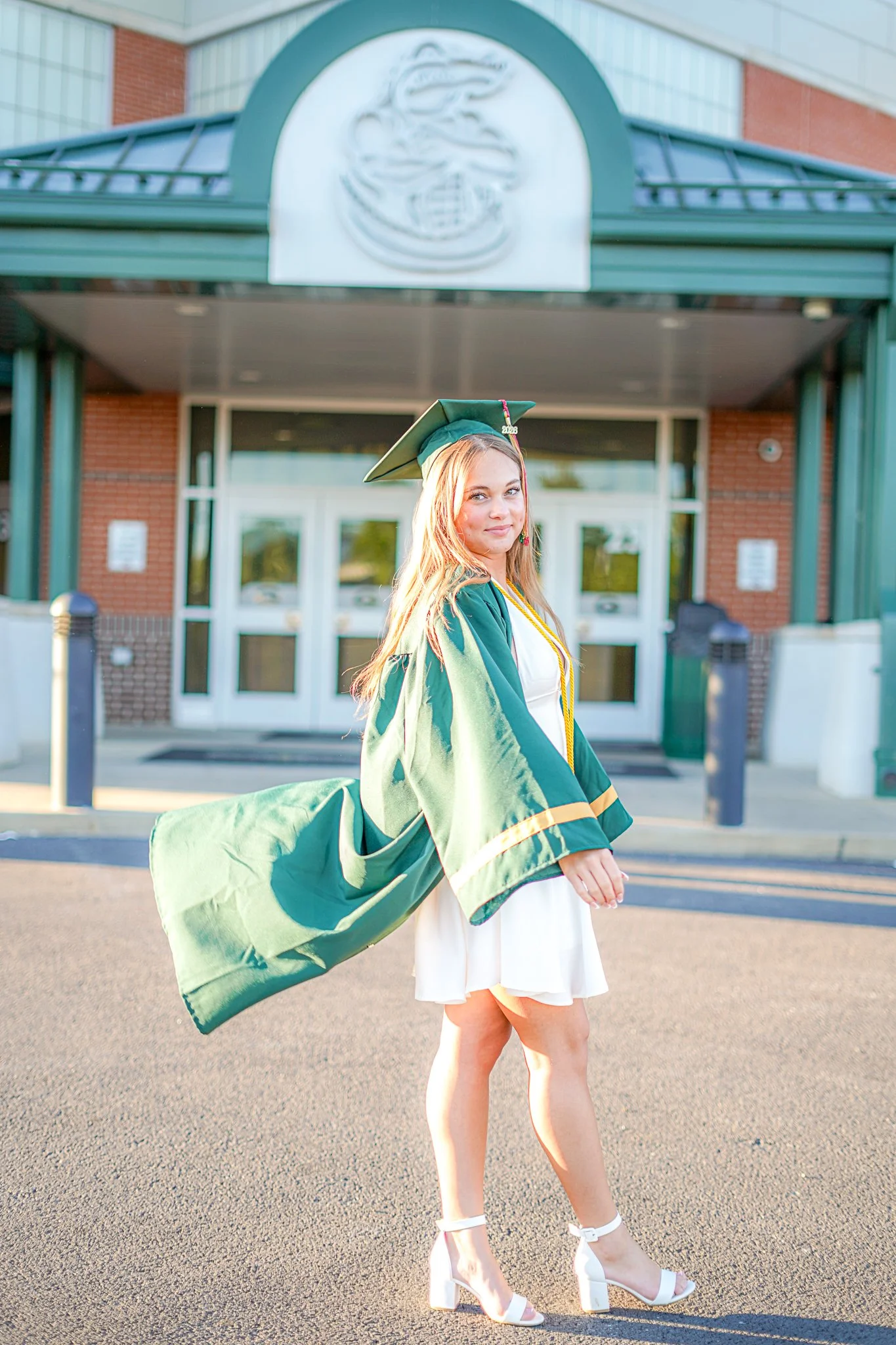 A young woman in a white dress and green graduation gown and cap standing outside in front of a school building, smiling at the camera, with her left hand on her side and her right arm slightly raised, wearing high-heeled white sandals.