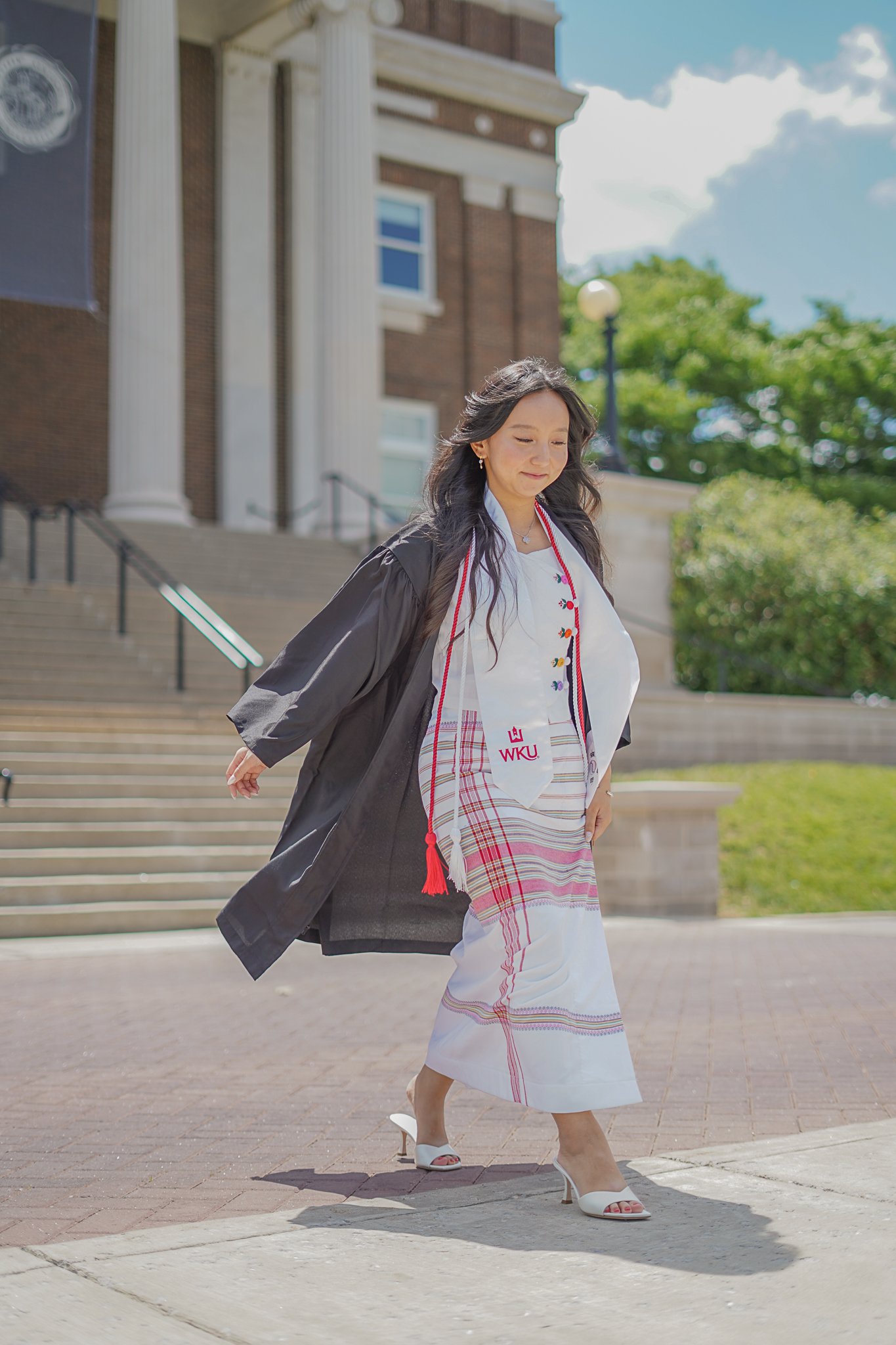 A woman wearing a graduation gown and a sash with the Western Kentucky University (WKU) logo walking down steps outdoors, smiling with her eyes closed. She is dressed in white heels and a white dress with a red and white plaid pattern. The background