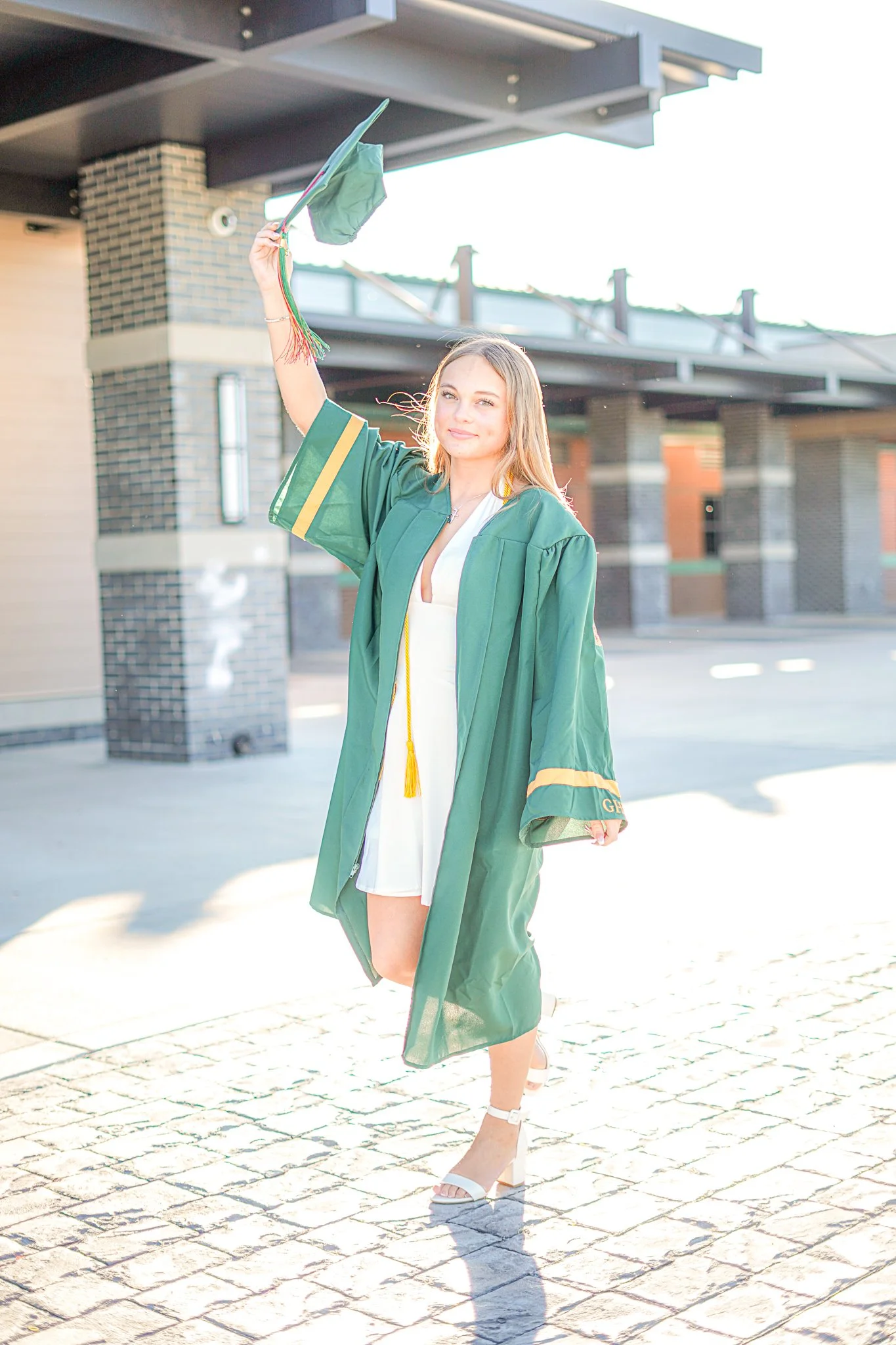 Young woman in a white dress and high heels celebrating graduation outdoors, wearing a green graduation gown and cap, raising her cap in the air with sunlight behind her.