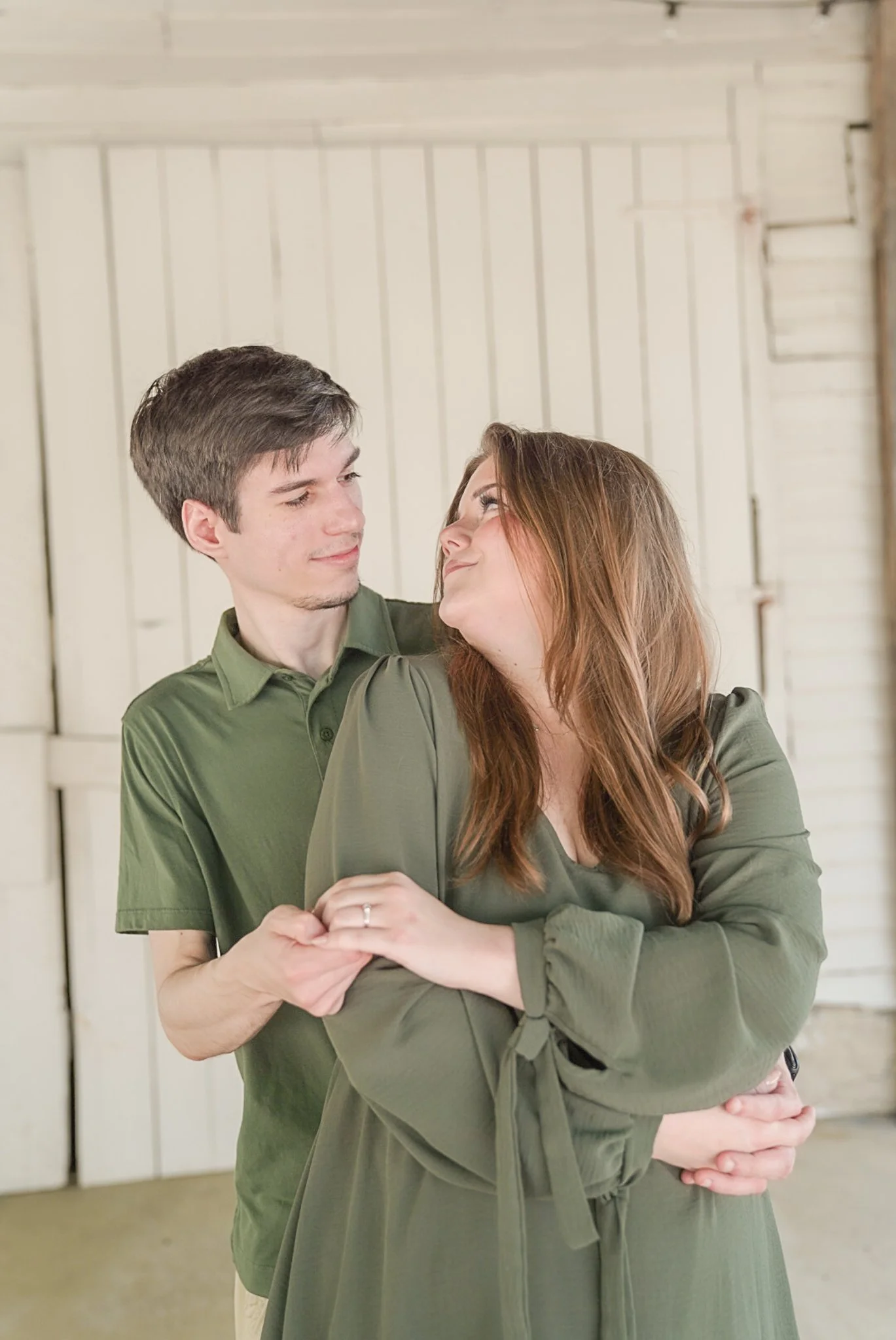 A couple holding hands and looking into each other's eyes indoors with a white wooden wall background.
