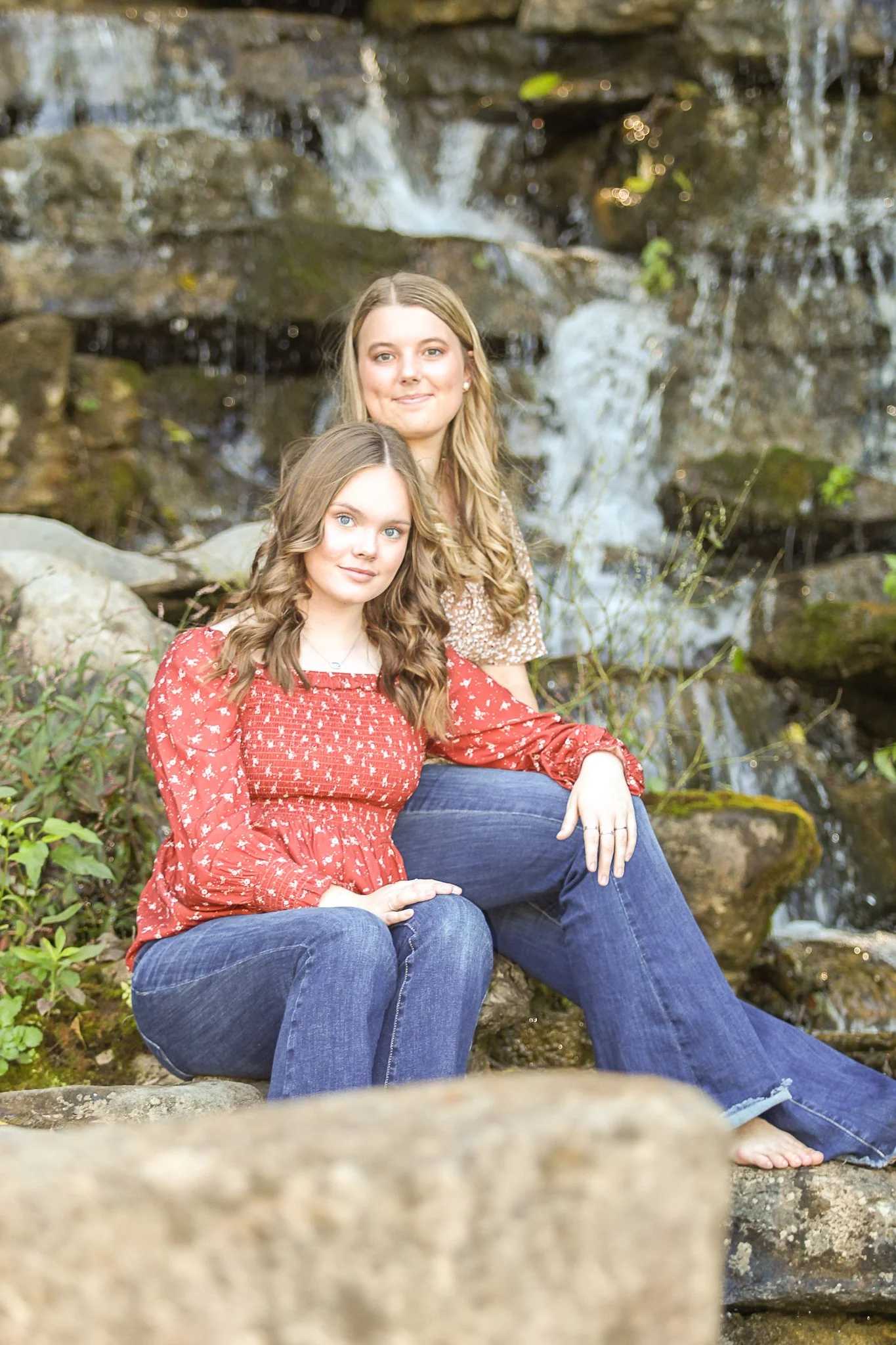 Two young women sitting by a small waterfall in a natural outdoor setting. The woman in the front is wearing a red patterned top and blue jeans, with long wavy brown hair and bright blue eyes. The woman in the back has long blonde hair, a light brown