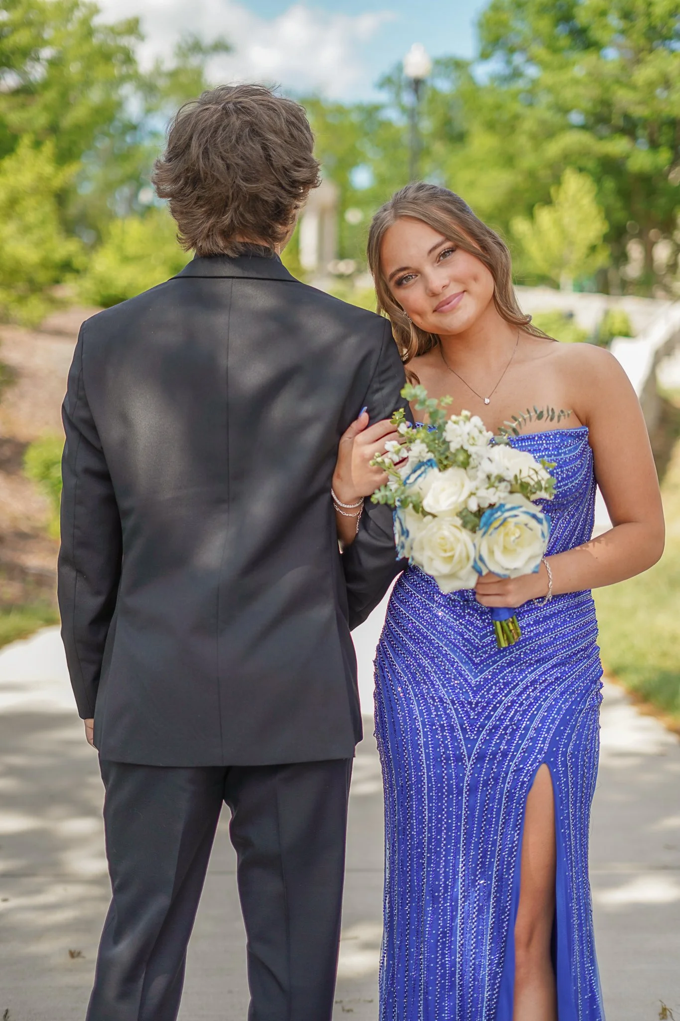 A woman in a blue sequined gown holding a bouquet of white roses, smiling, with a man in a black tuxedo, in an outdoor park setting.