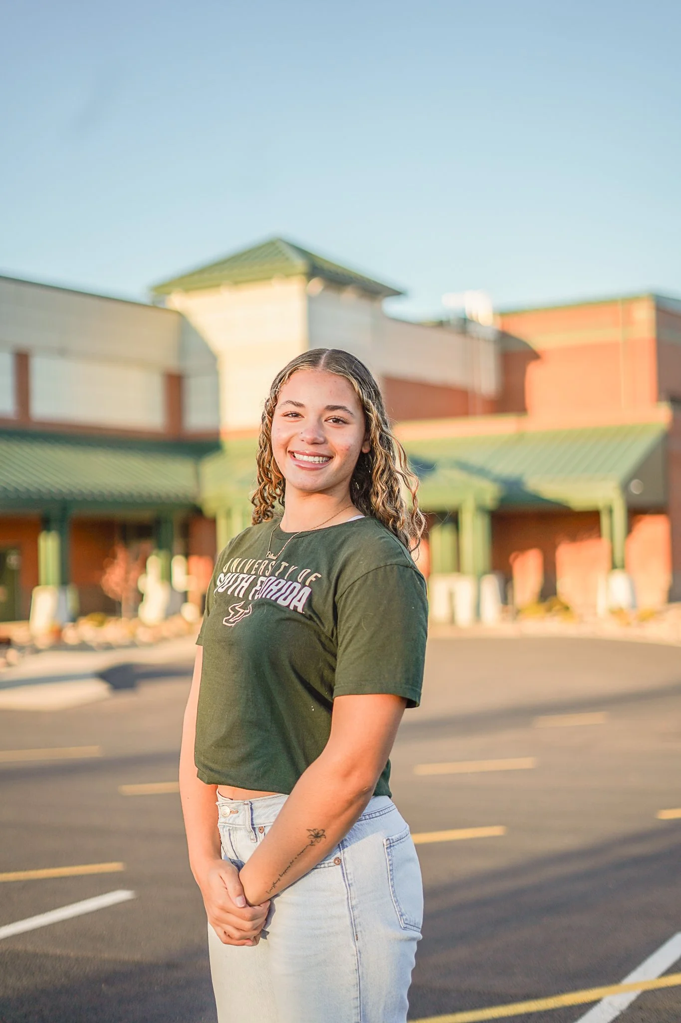 A young woman with curly hair smiling outdoors in a parking lot with a commercial building behind her, wearing a green T-shirt and light-colored jeans.