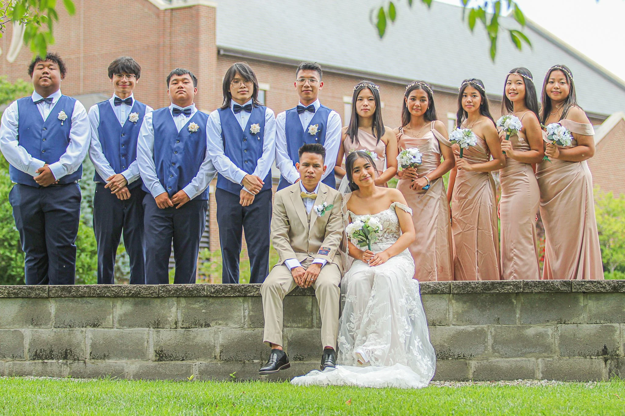 A group of ten young people, six men and four women, dressed in formal attire, standing outdoors on a brick structure. Two women in wedding dresses are seated in front, with one man in a beige suit and yellow bowtie seated beside them. The rest are s