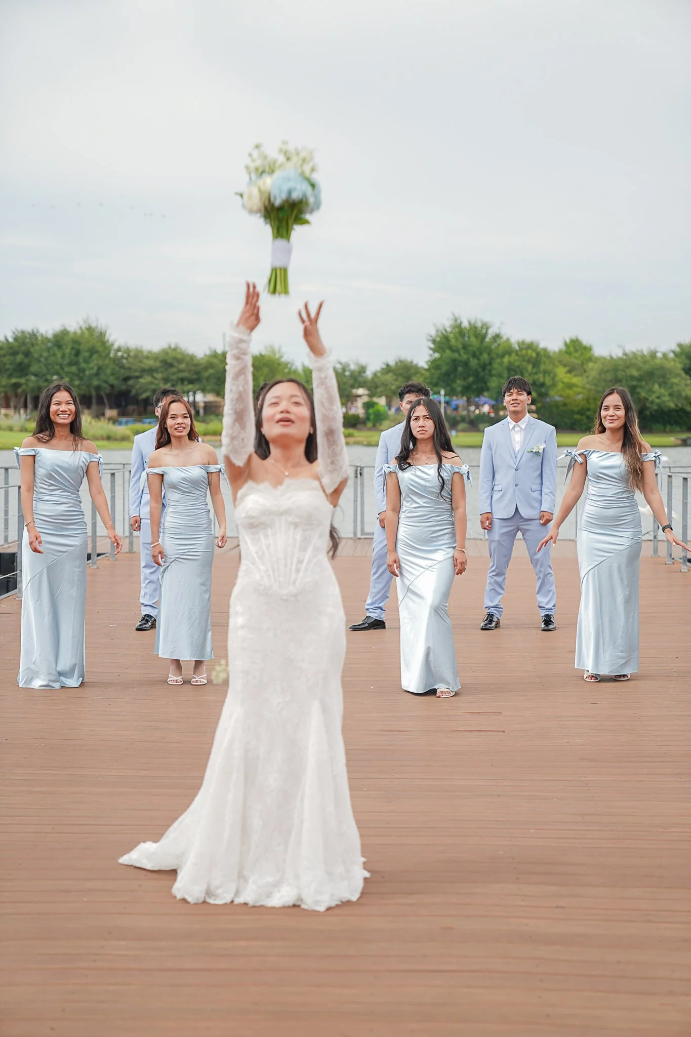 A bride in a white wedding dress catches a bouquet of flowers during an outdoor wedding ceremony, with six bridesmaids and a groom standing behind her on a wooden deck near a body of water, surrounded by green trees.
