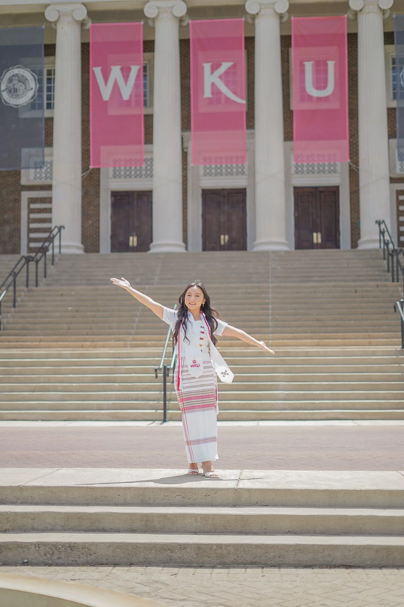 A young woman smiling and posing with arms outstretched in front of campus steps, in front of a building with Revolutionary War era style larger white columns. There are large pink banners with white letters spelling WKU hanging between the columns. 