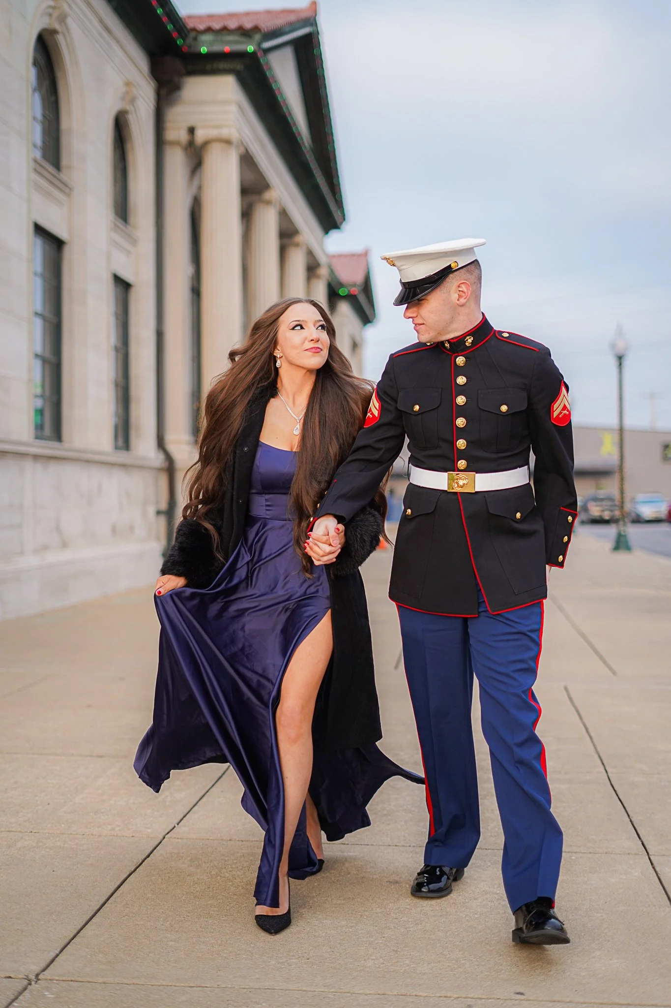 A woman in an elegant dark purple dress walking hand in hand with a Marine in dress uniform on a city sidewalk.