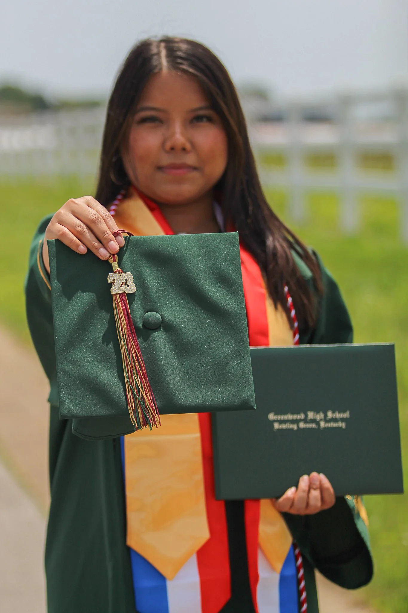 A woman in graduation cap and gown holding a green diploma cover and a tassel with a 23 charm. She is outdoors, with a blurred background, and is wearing a graduation stole with American flag colors.