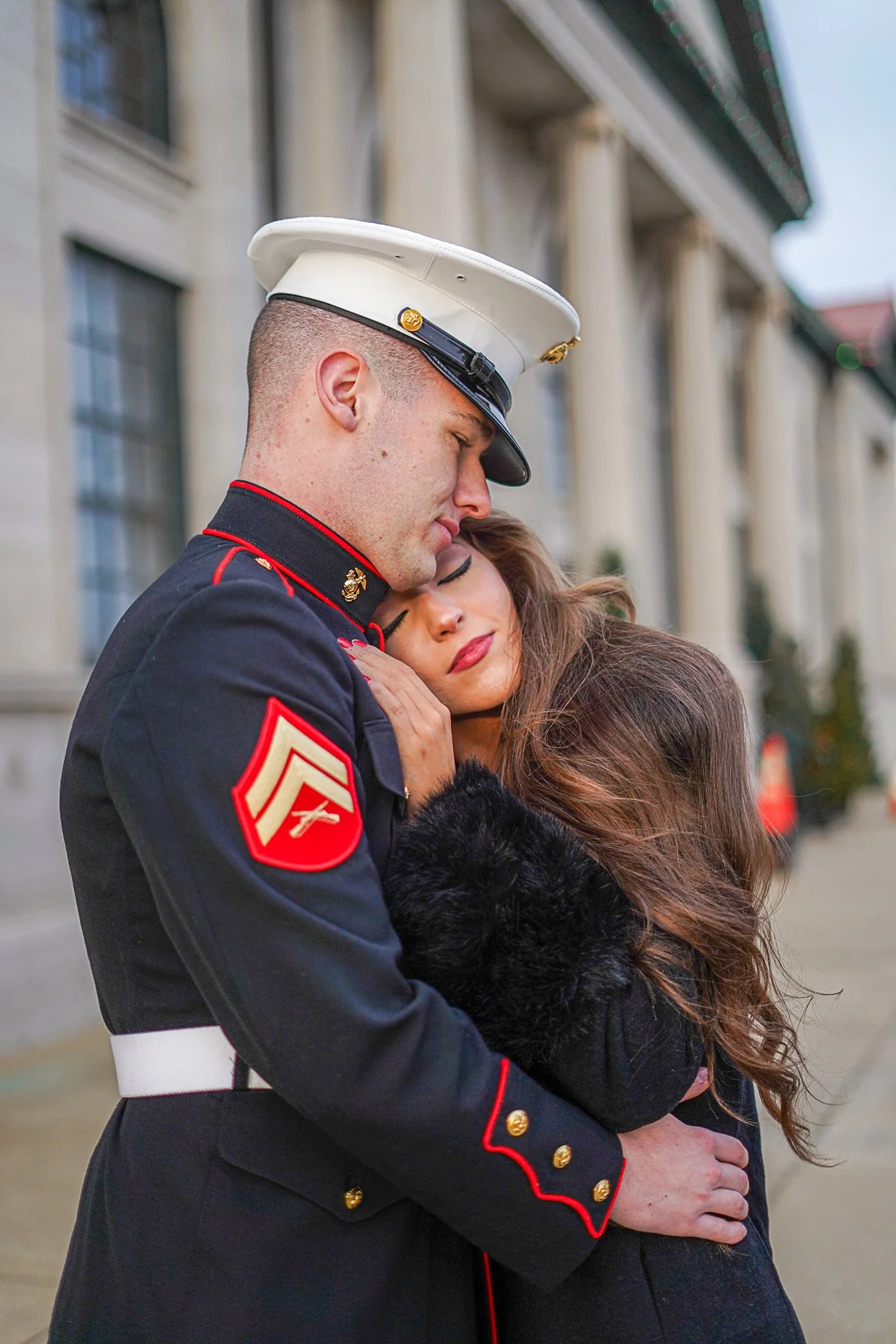 A Marine in uniform embraces a woman, both with their eyes closed, outside a building with large columns and windows.