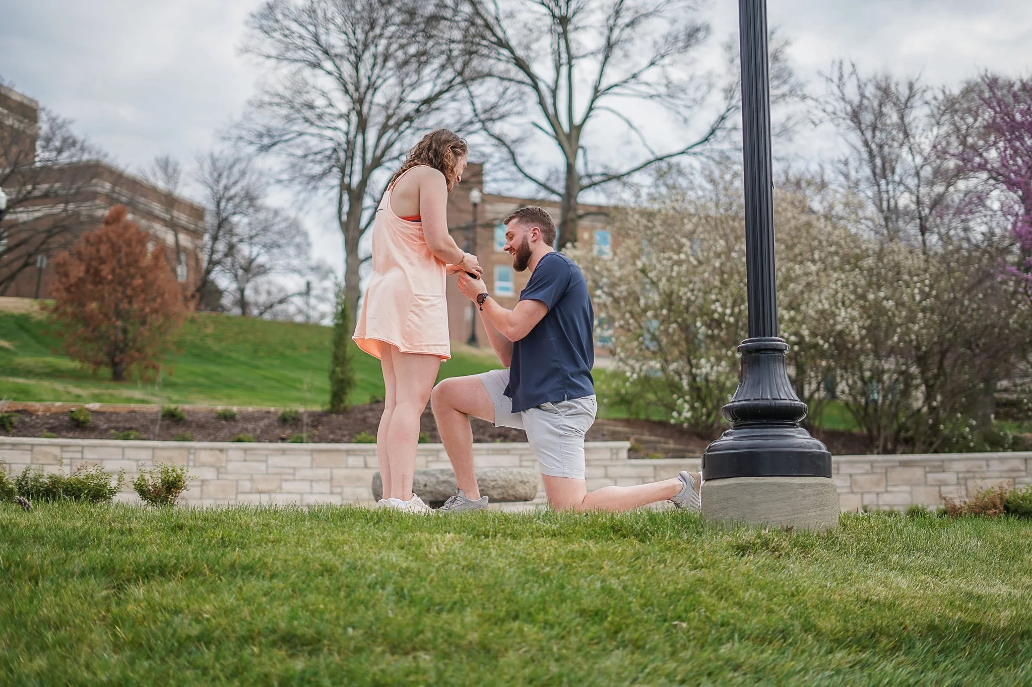 A man kneeling on one knee proposing marriage to a woman in a park with trees and buildings in the background.