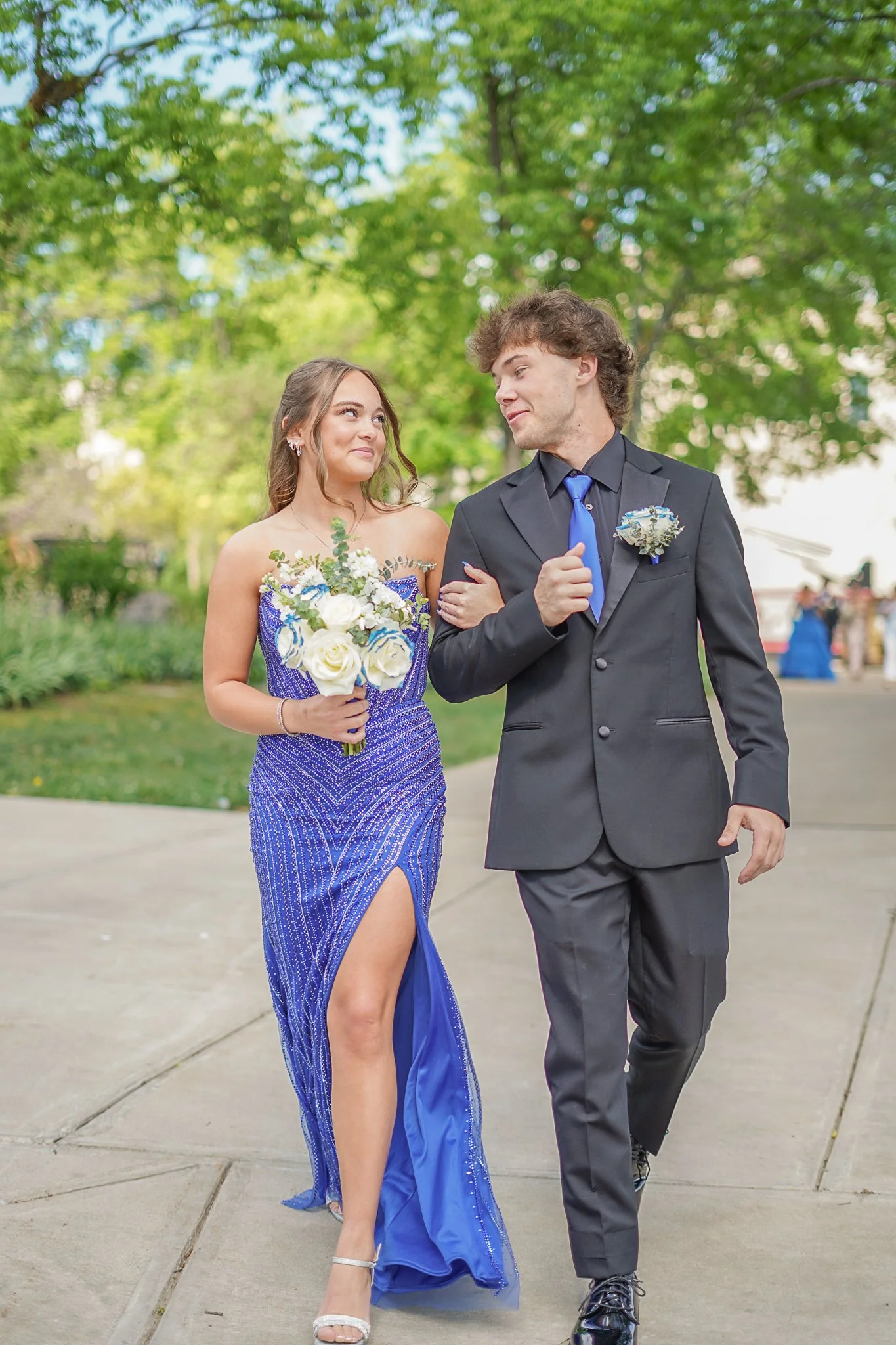 A young woman in a blue evening gown holding a bouquet of white flowers, walking arm-in-arm with a young man in a gray suit with a blue tie, both smiling and looking at each other outdoors with green trees in the background.