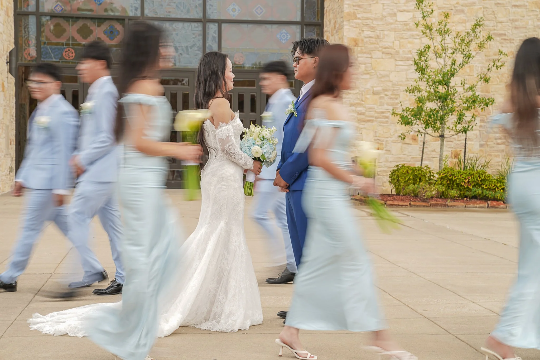 A bride and groom holding hands during their wedding ceremony, with bridesmaids and groomsmen walking past in motion blur.