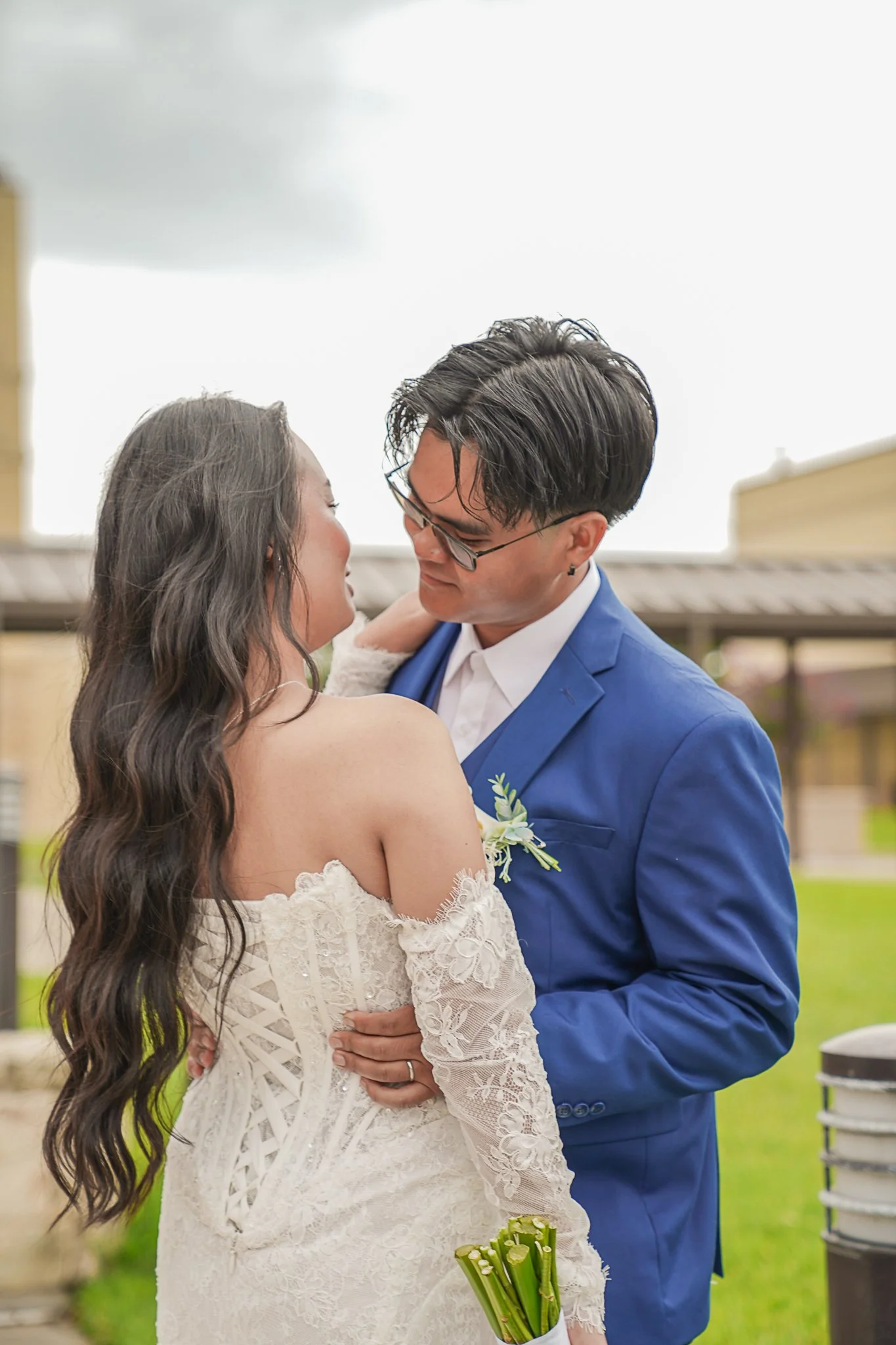 A bride and groom embrace outdoors on a cloudy day. The bride wears a white lace wedding dress with long sleeves and has long wavy dark hair. The groom wears a blue suit with a white shirt and has dark hair styled back, wearing glasses. The bride hol