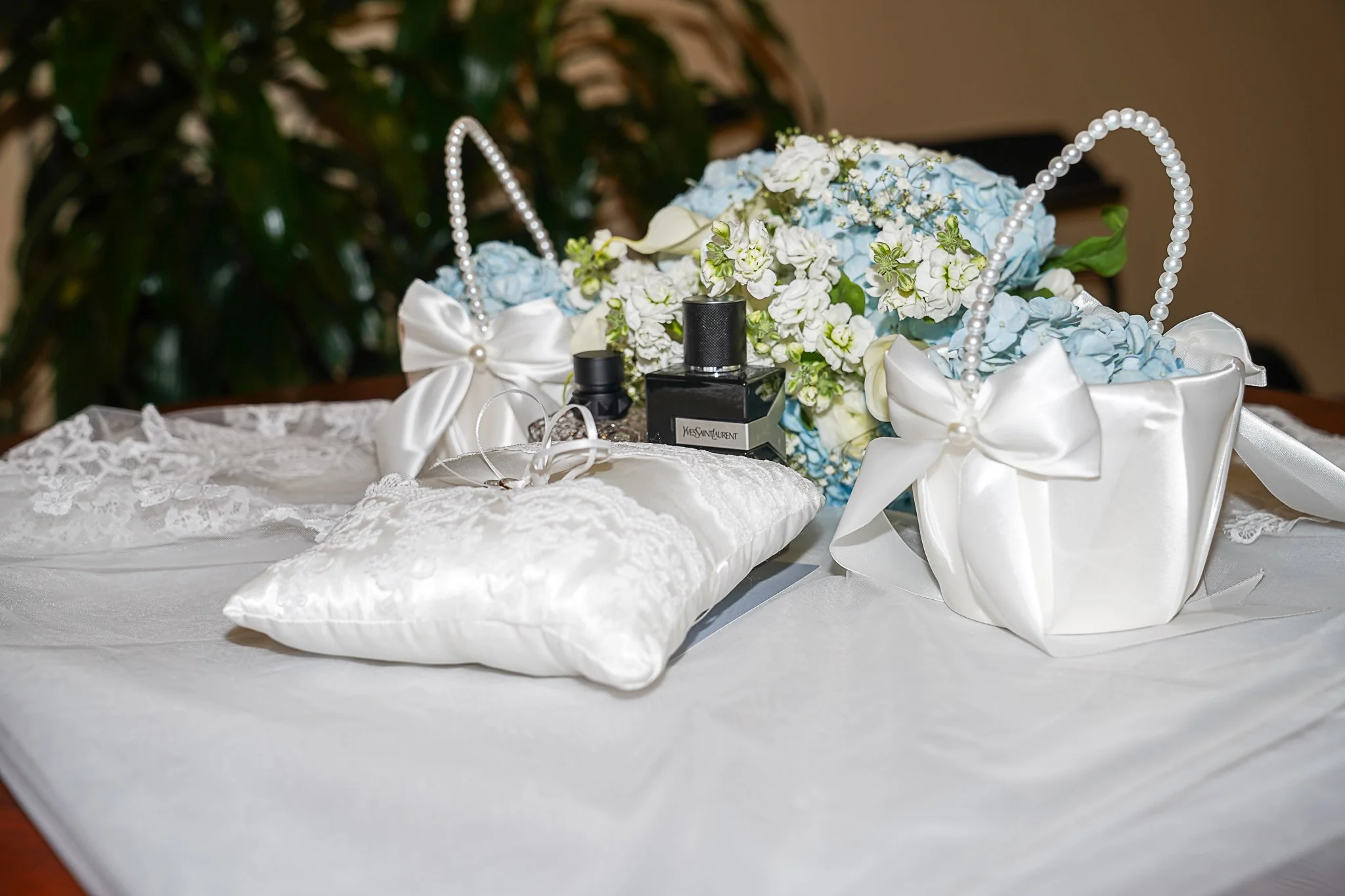 A decorative wedding arrangement with a floral bouquet in shades of white and blue, white satin bows, pearl accents, a small black Yves Saint Laurent perfume bottle, and a white lace pillow on a white tablecloth.