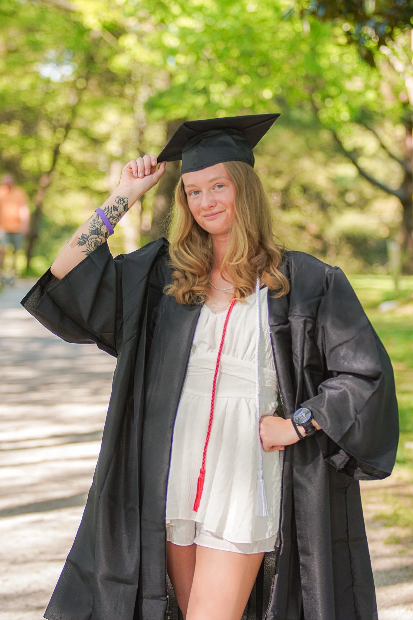 Young woman in a graduation gown and cap outdoors, holding her cap with her right hand, smiling, with trees in the background.