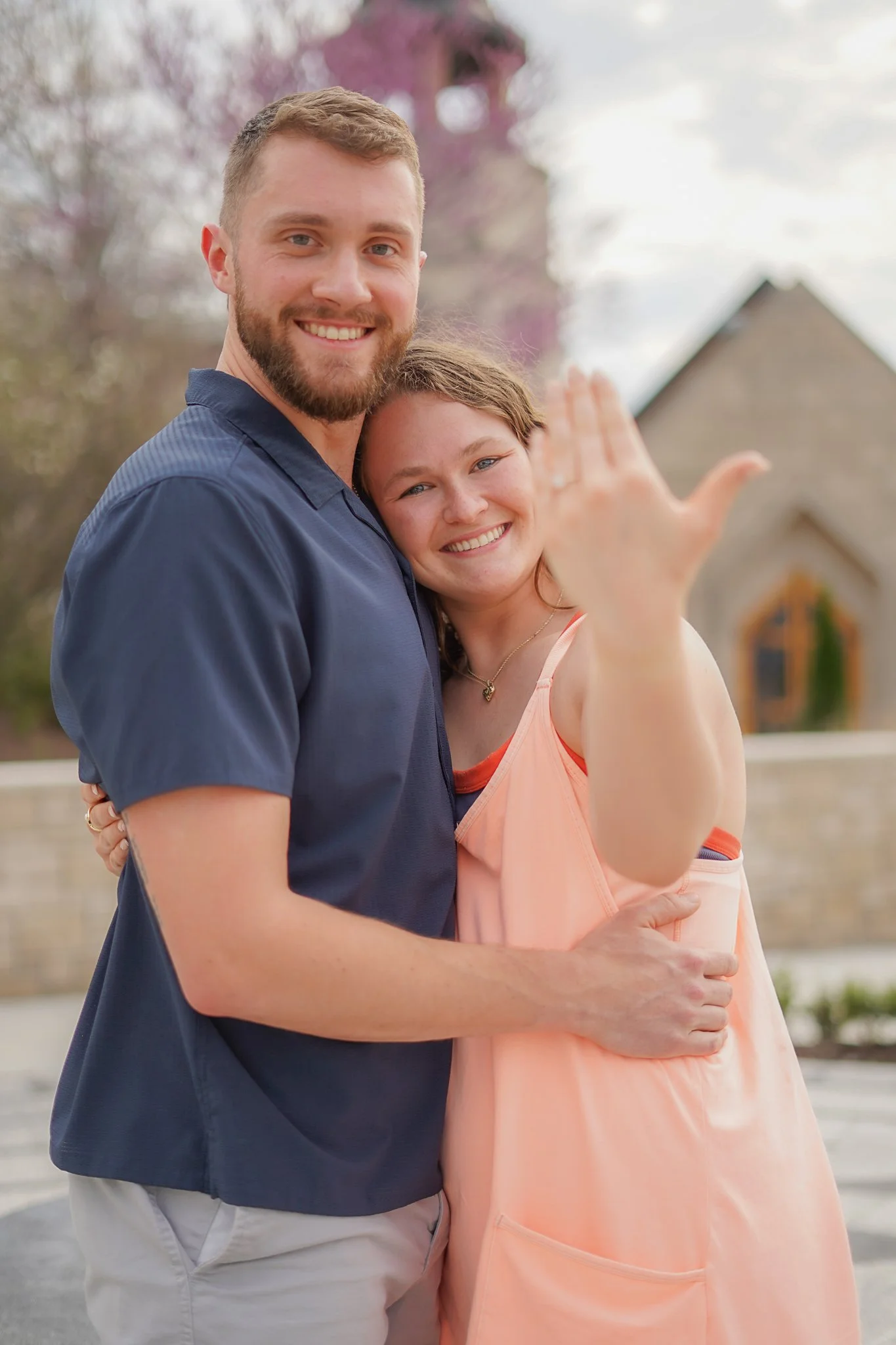 A young man and woman smiling and embracing outdoors, with the woman waving her hand.