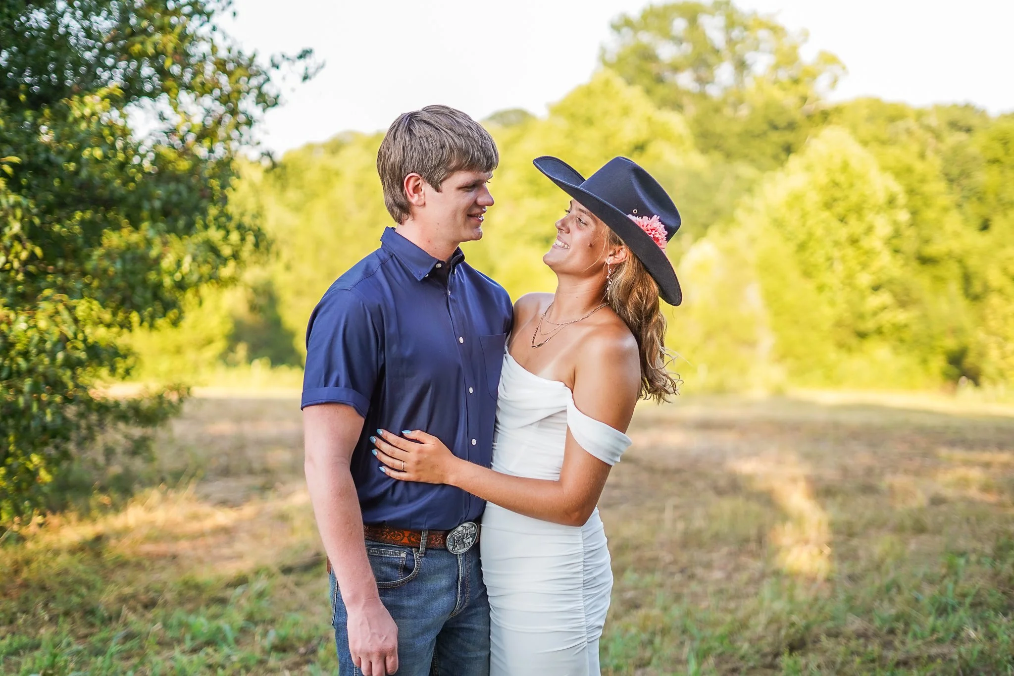 A couple stands close together outdoors in a park, gazing into each other's eyes, with trees and sunlight in the background.
