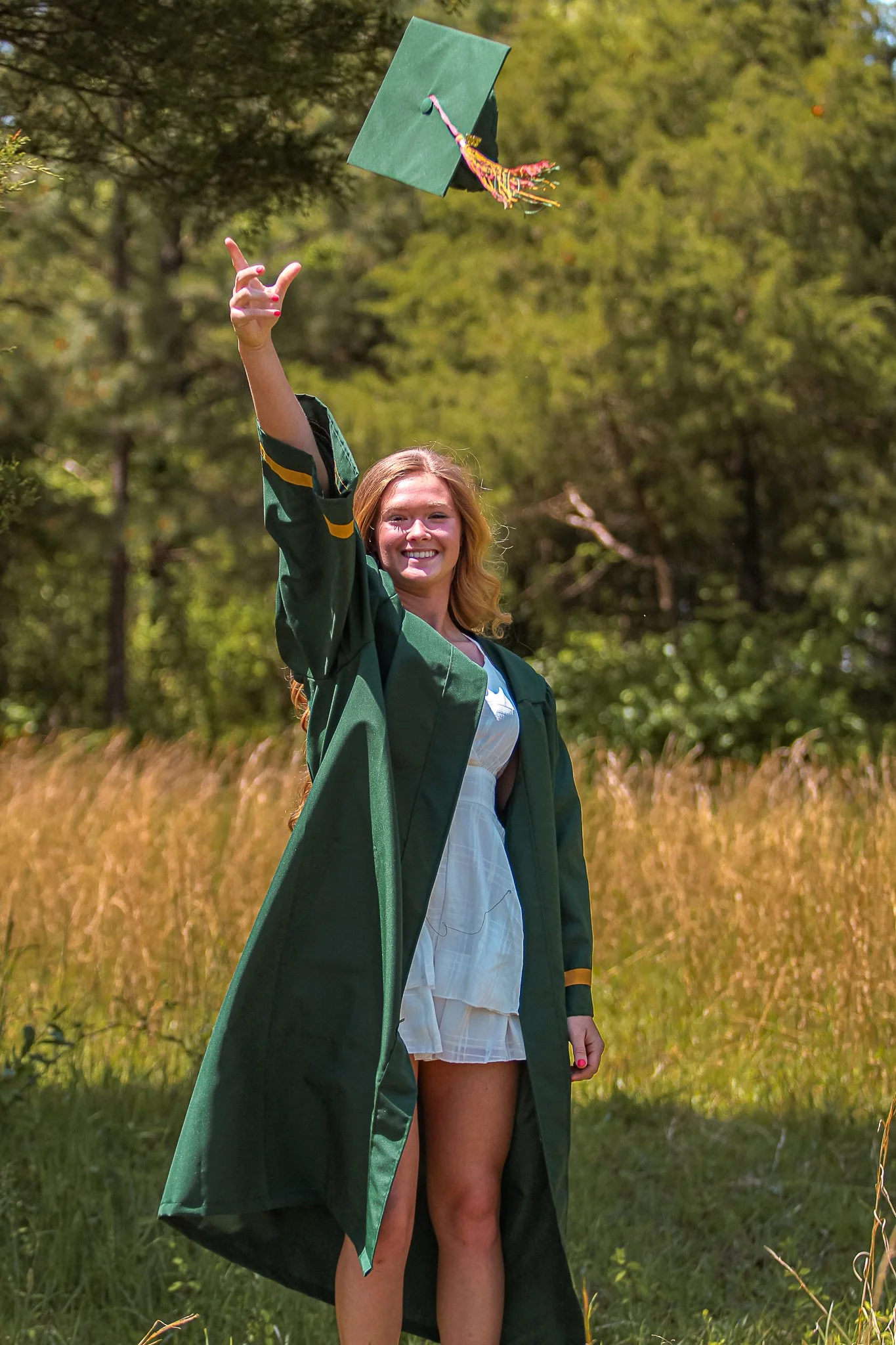 A woman in a green graduation gown is outdoors, throwing her green graduation cap in the air while smiling. She is standing in a field with tall grass and trees in the background.