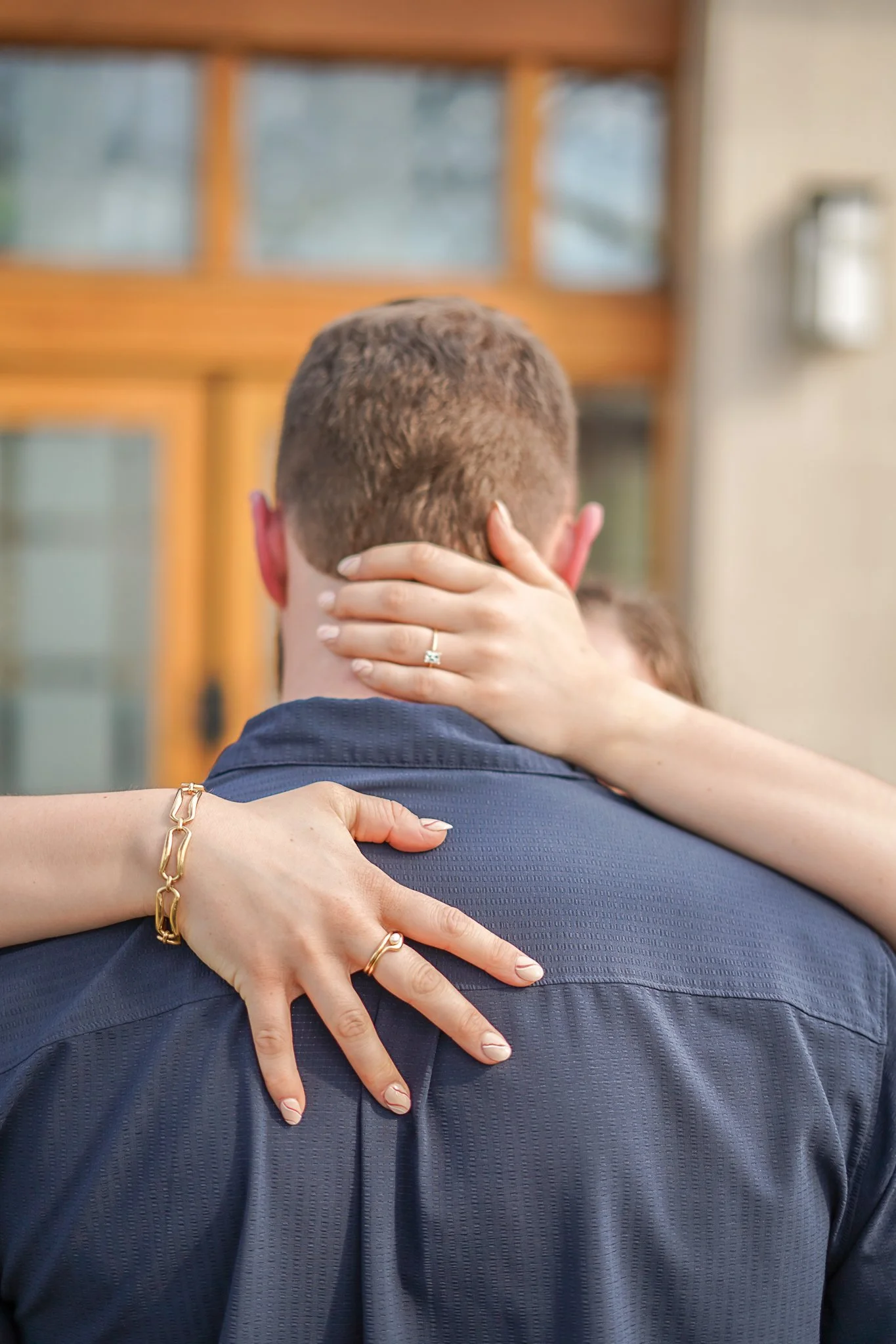 A couple dancing closely, the woman's hands around the man's neck, and her face partially visible as she leans in towards him. The man is seen from behind, wearing a dark blue shirt, with a wooden structure with windows in the background.