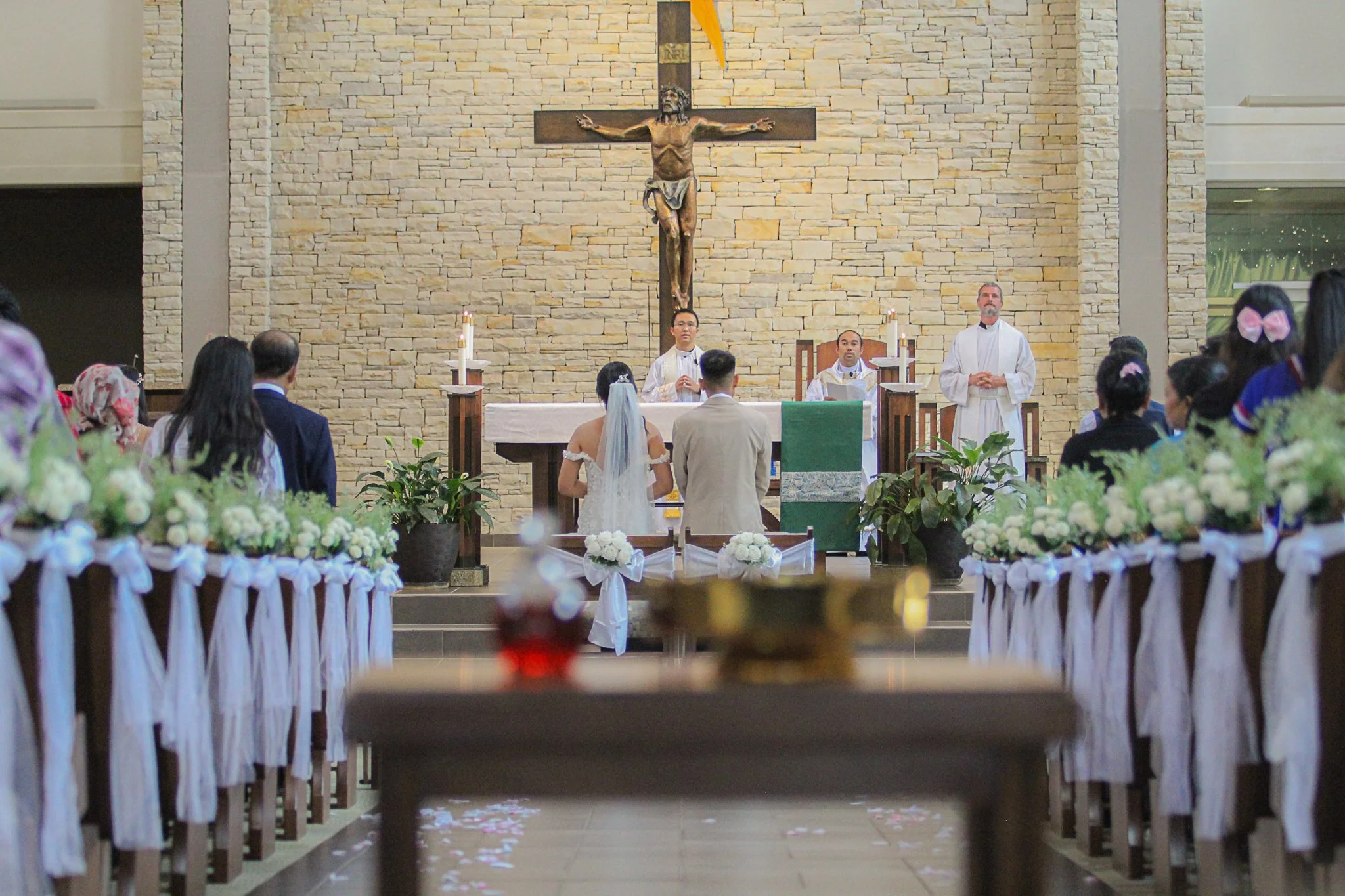 A wedding ceremony taking place inside a church with a congregation seated in pews decorated with white ribbons and flowers. The bride and groom stand at the altar facing a priest and two other clergy members. Behind them is a large crucifix on a sto