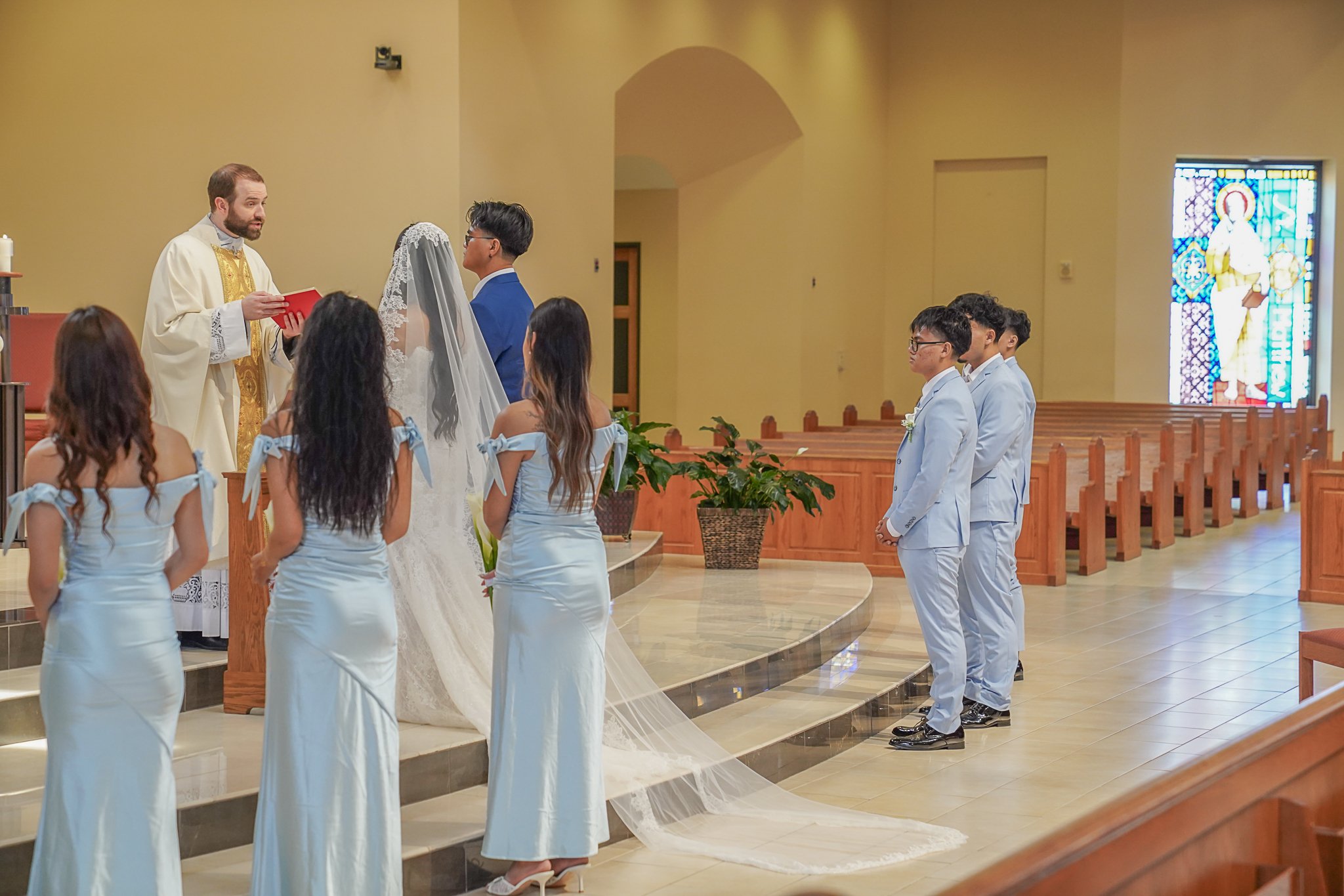 A wedding ceremony taking place in a church with couples standing in front of a priest during the vows. The bride is wearing a white wedding dress and veil, and the groom is dressed in a blue suit. The bridesmaids are in light blue dresses, and the g