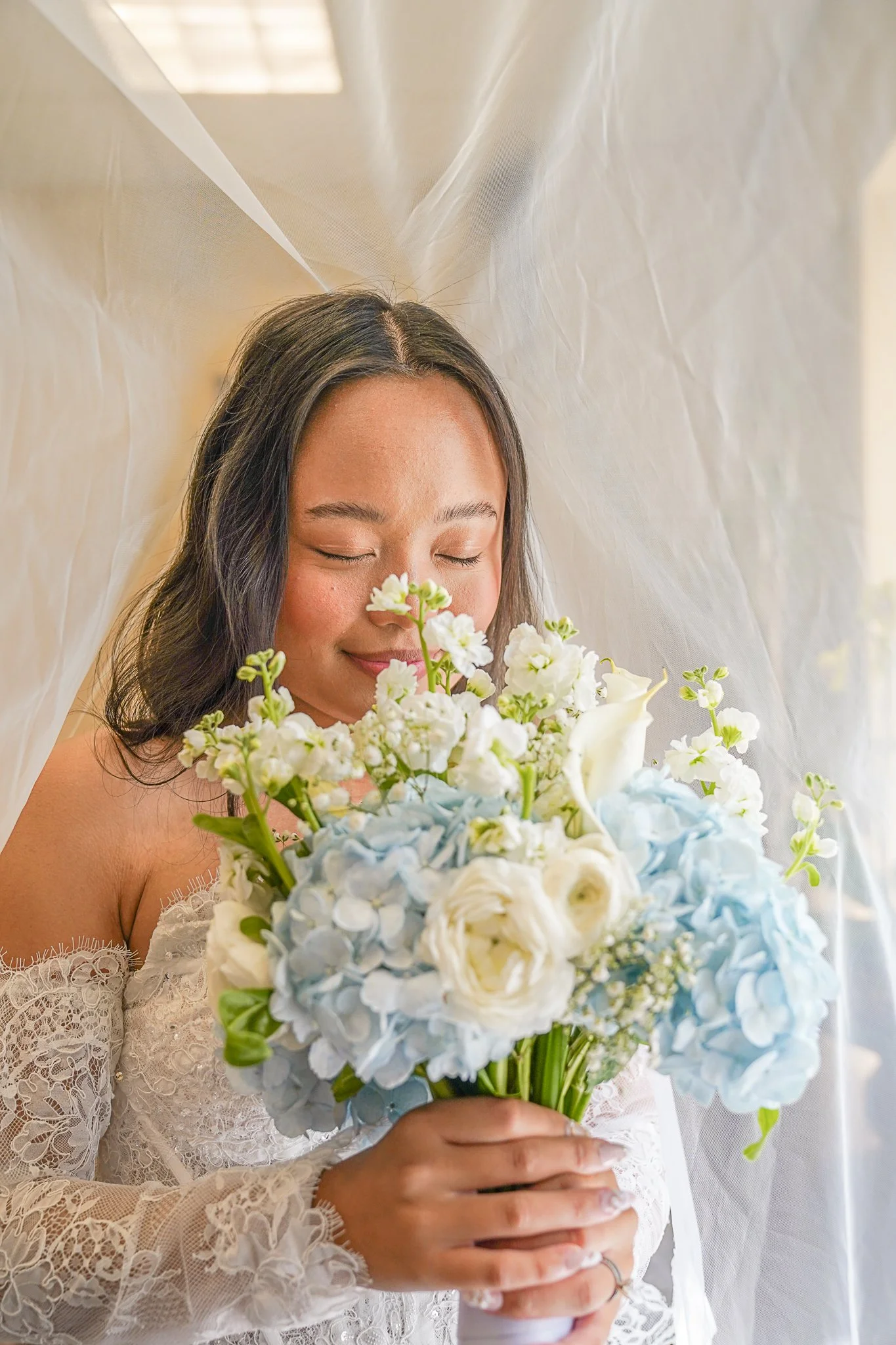 A woman in a white lace wedding dress holding a bouquet of white and light blue flowers, with her eyes closed and a soft smile.