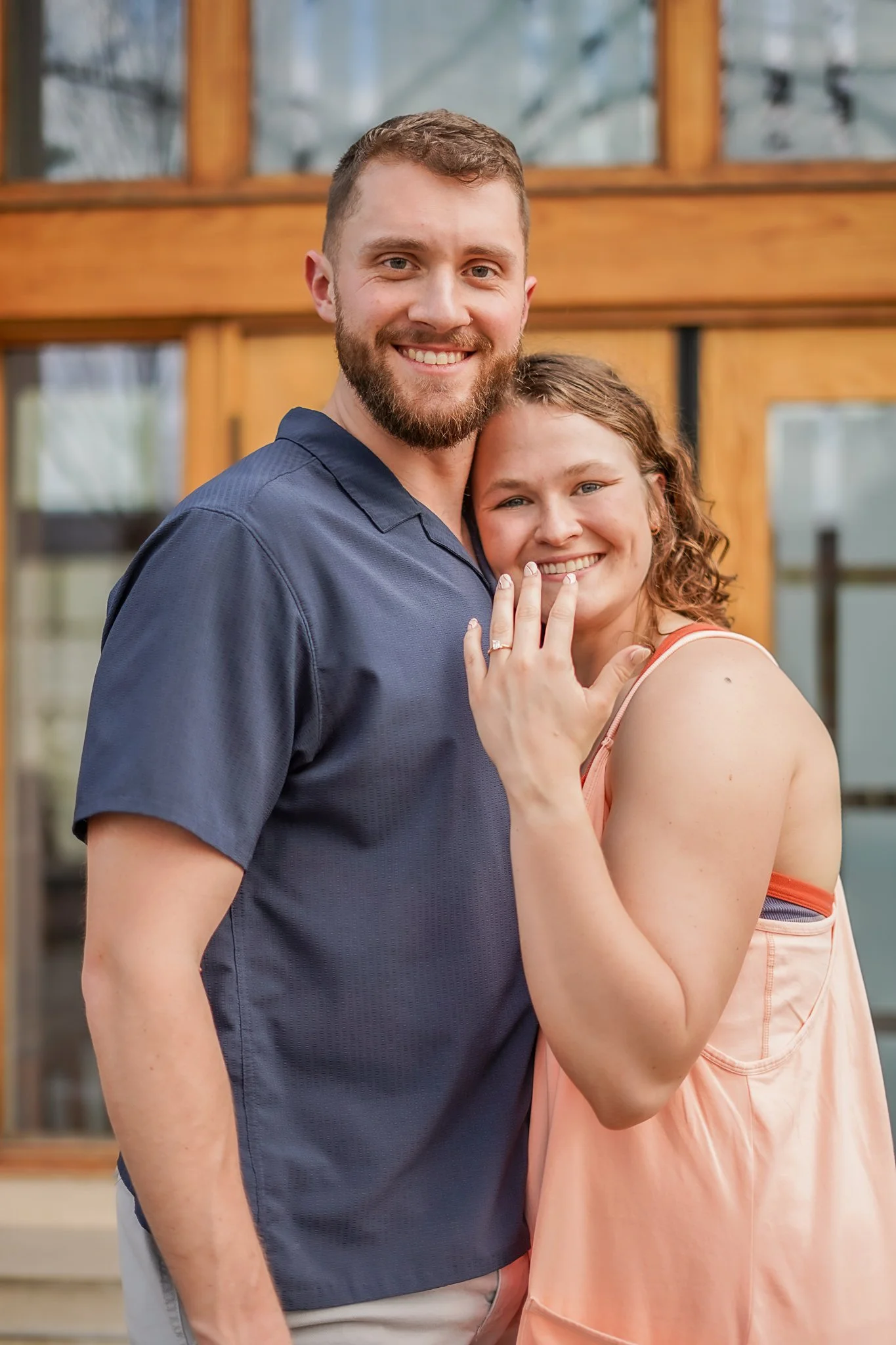 A happy man with a beard and a woman with curly hair smiling and hugging outside near a wooden building with glass windows.