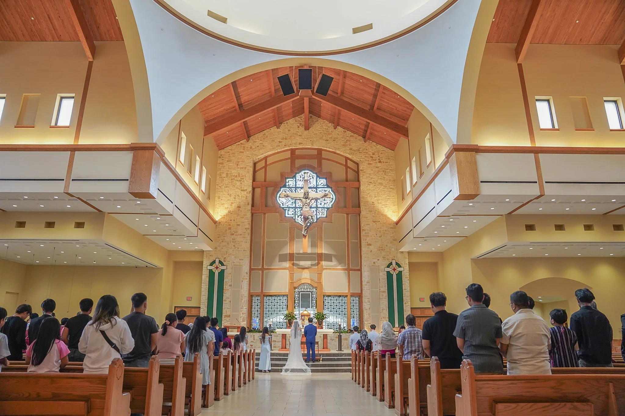 Inside a church during a wedding ceremony with a congregation facing the altar, which features a crucifix and stained glass window, with wooden pews and high ceiling.