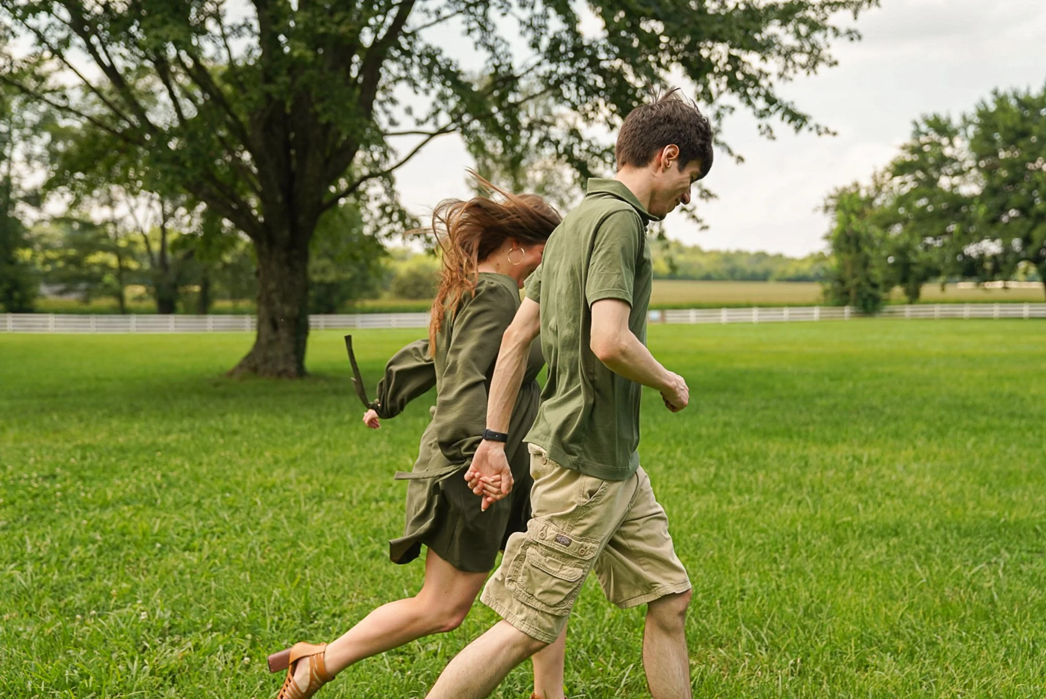 A couple holding hands and running on a grassy field near a large tree and a white fence in a rural area on a cloudy day.
