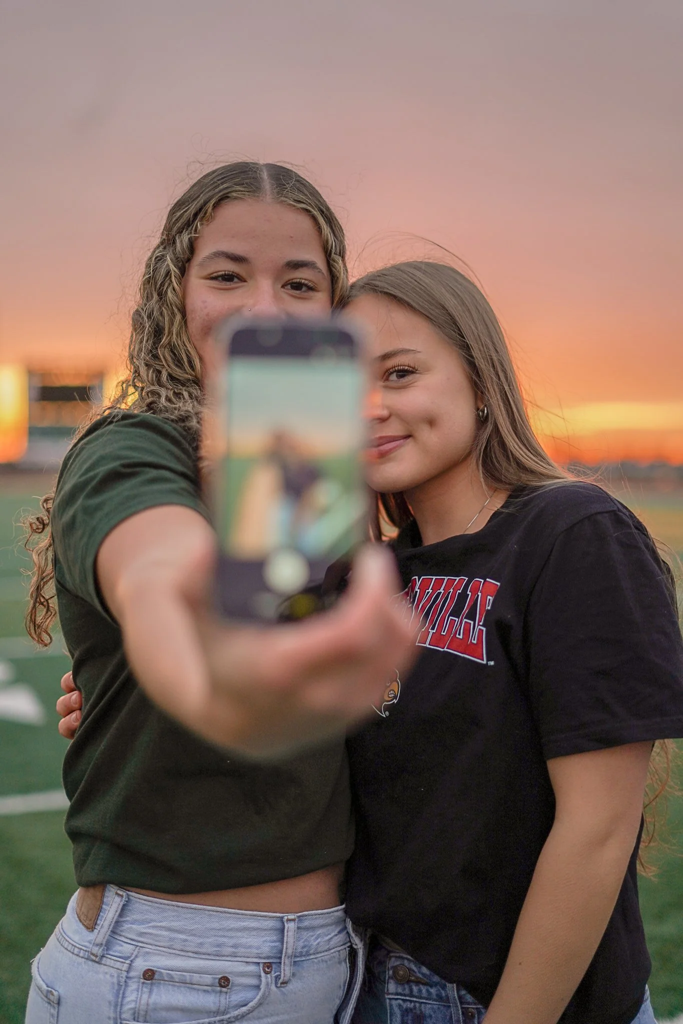 Two young women taking a selfie outdoors during sunset. One woman with curly hair and a nose piercing holds the phone, partially visible in the foreground. The other woman with straight hair leans close, smiling. The background features a colorful sk