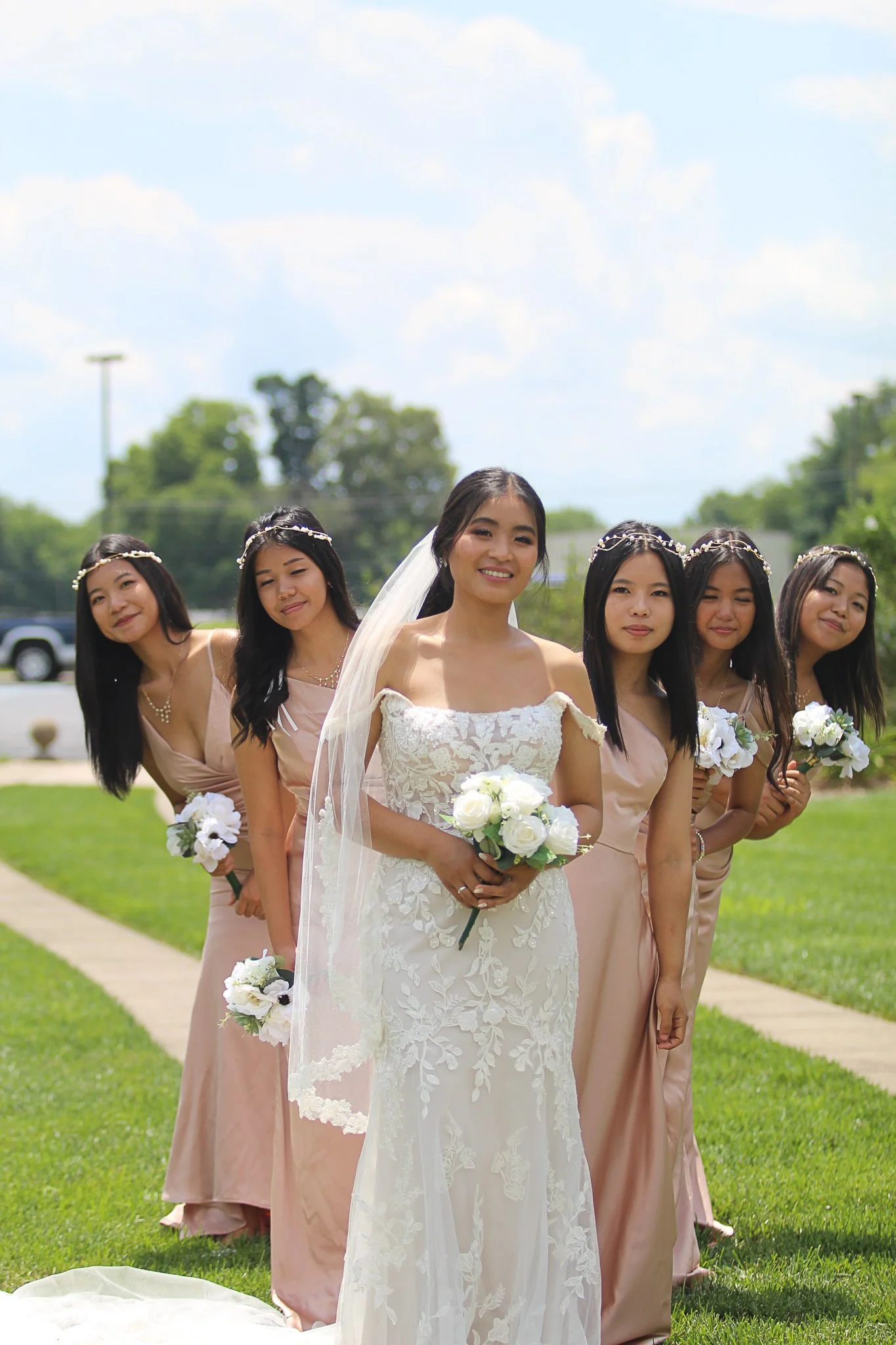 A bride in a white lace wedding dress and veil holding a bouquet of white roses, standing outdoors with five bridesmaids in matching blush pink dresses, all wearing headbands and holding bouquets, on a grassy area with trees and a partly cloudy sky i