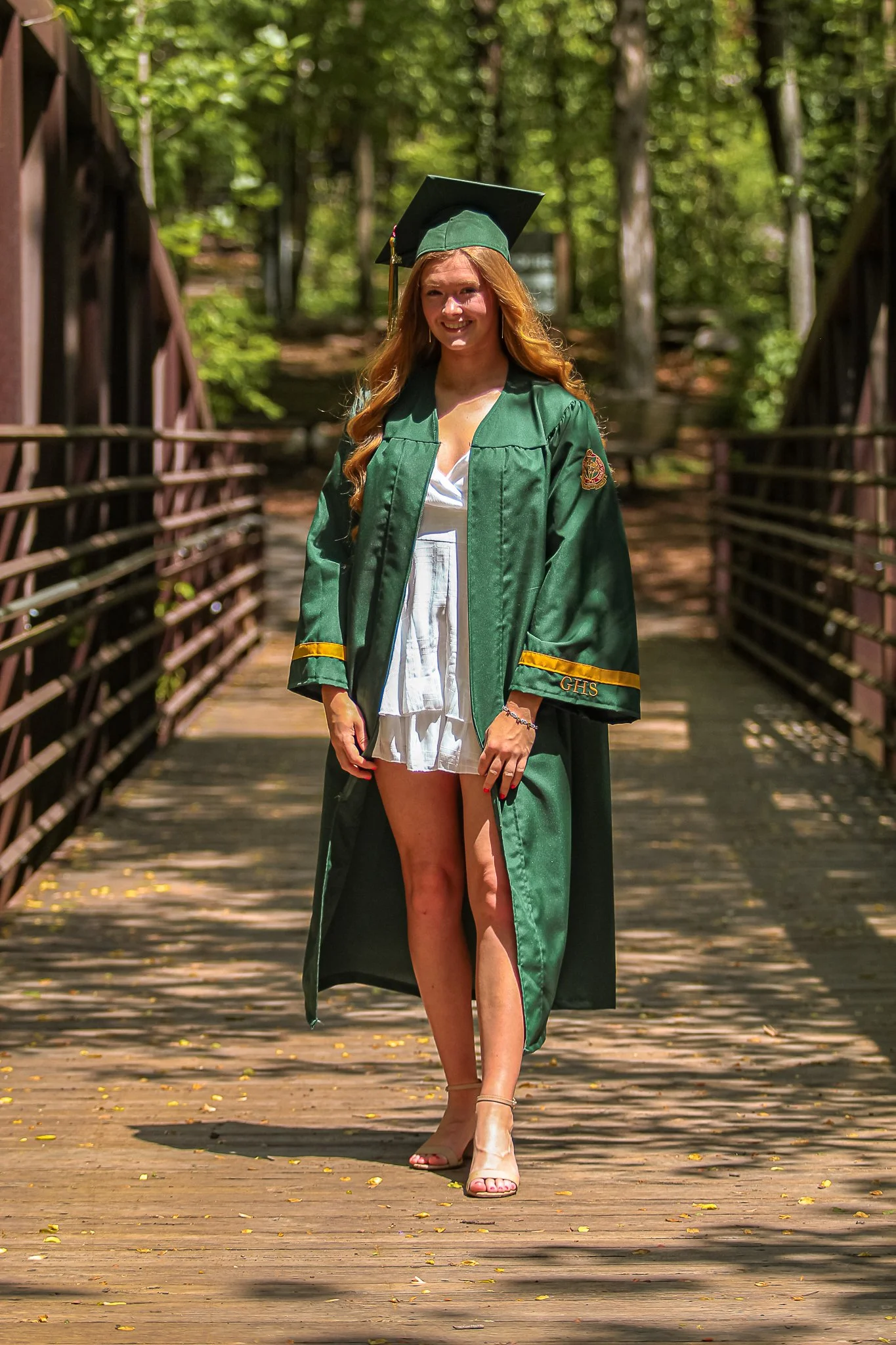 A woman in a green graduation gown and cap standing on a wooden bridge in a wooded area. She is smiling and wearing high heels.