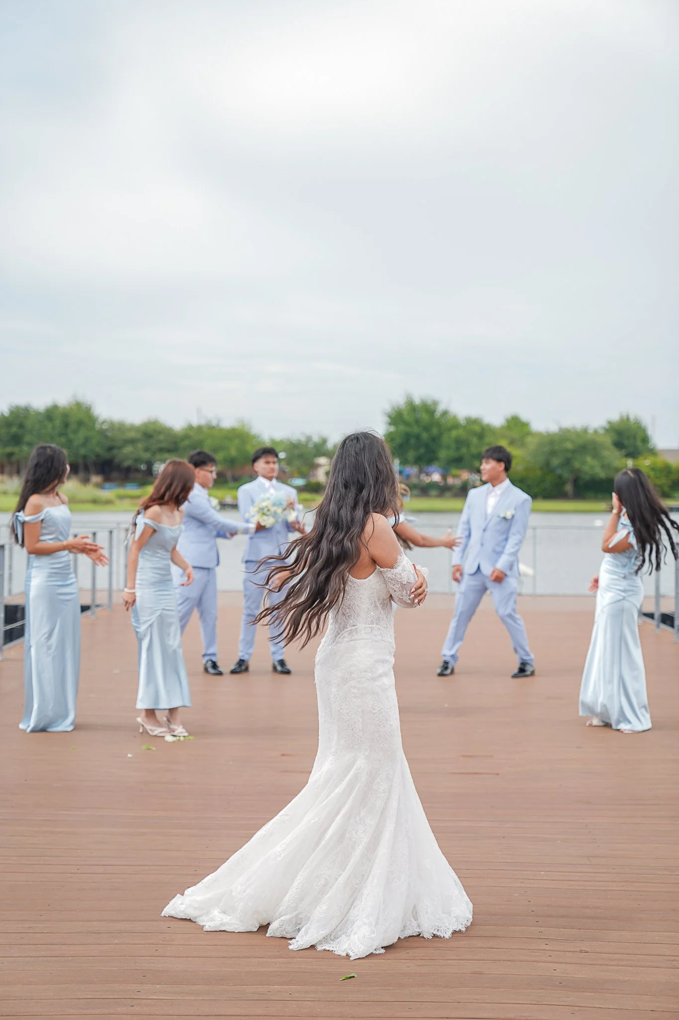 A bride in a white lace wedding dress standing with her back to a group of people outdoors during a wedding ceremony. The group includes bridesmaids in light blue dresses and groomsmen in light gray suits.