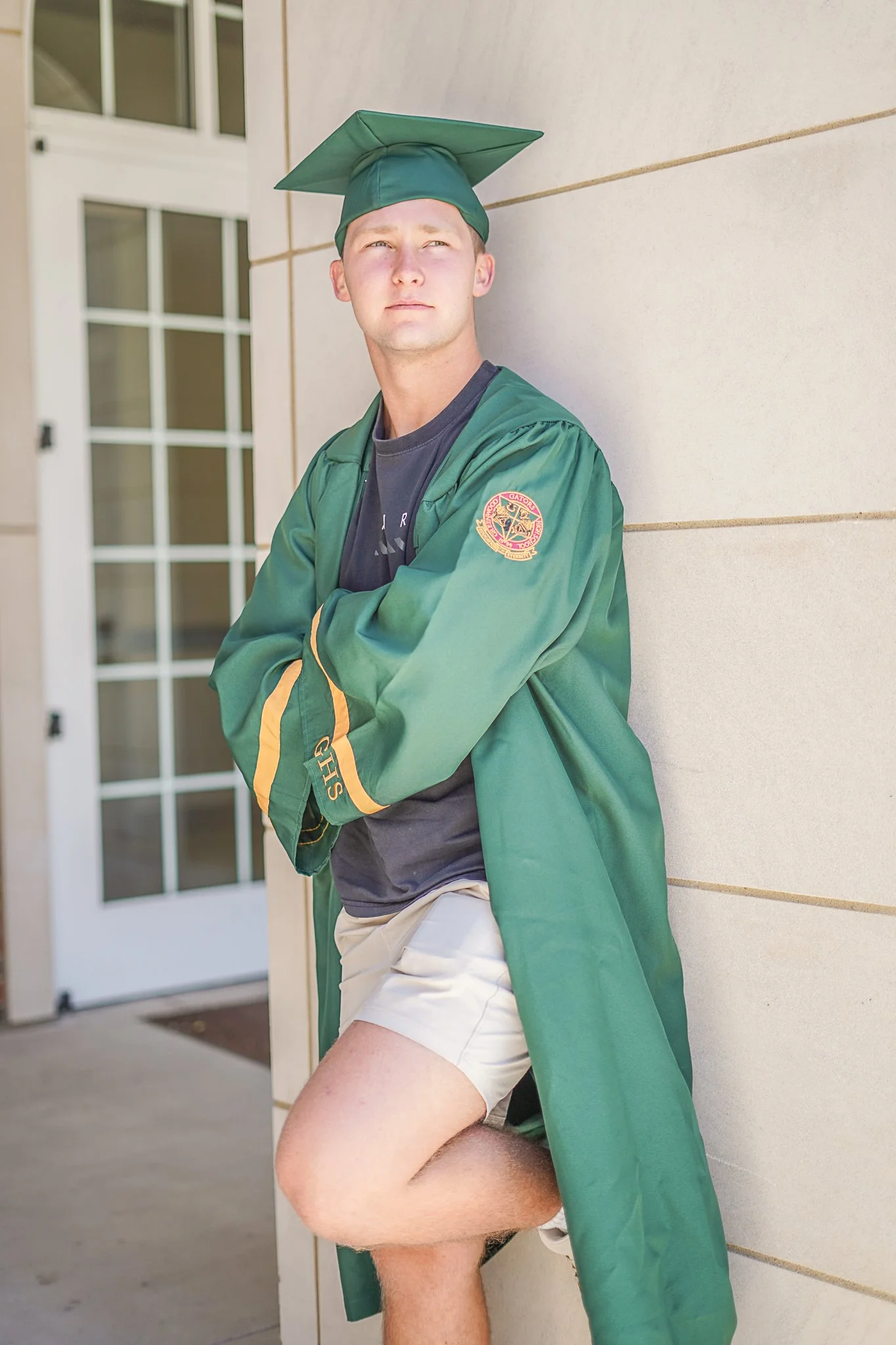 A young man in a green graduation gown and cap with a tassel, wearing a dark t-shirt and white shorts, leaning against a wall outside a building with glass doors, with his arms crossed and looking into the distance.