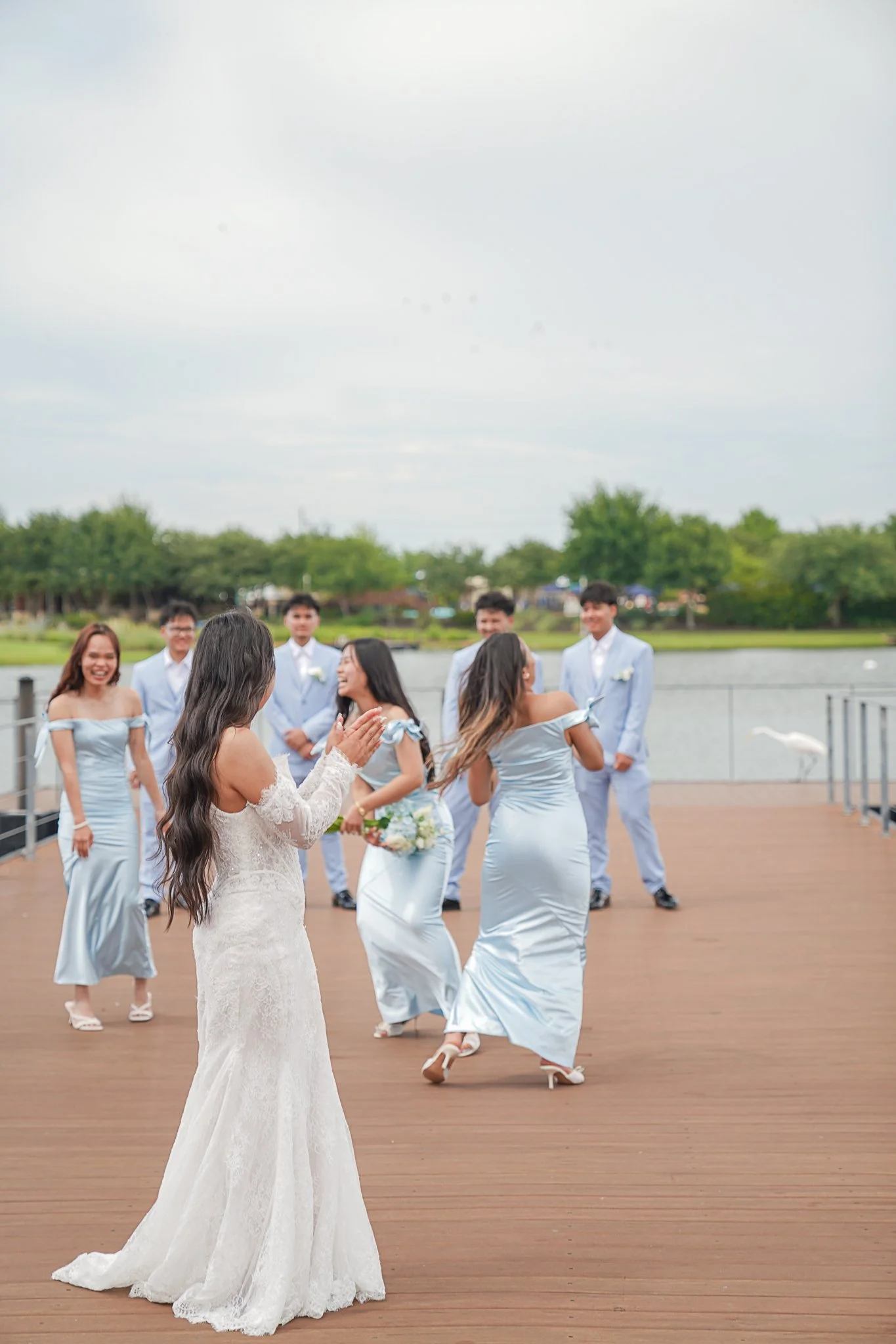 A bride in a white lace wedding dress with long dark hair stands on a wooden deck, facing a group of bridesmaids and groomsmen outside by a river with trees in the background, during a wedding ceremony or reception.