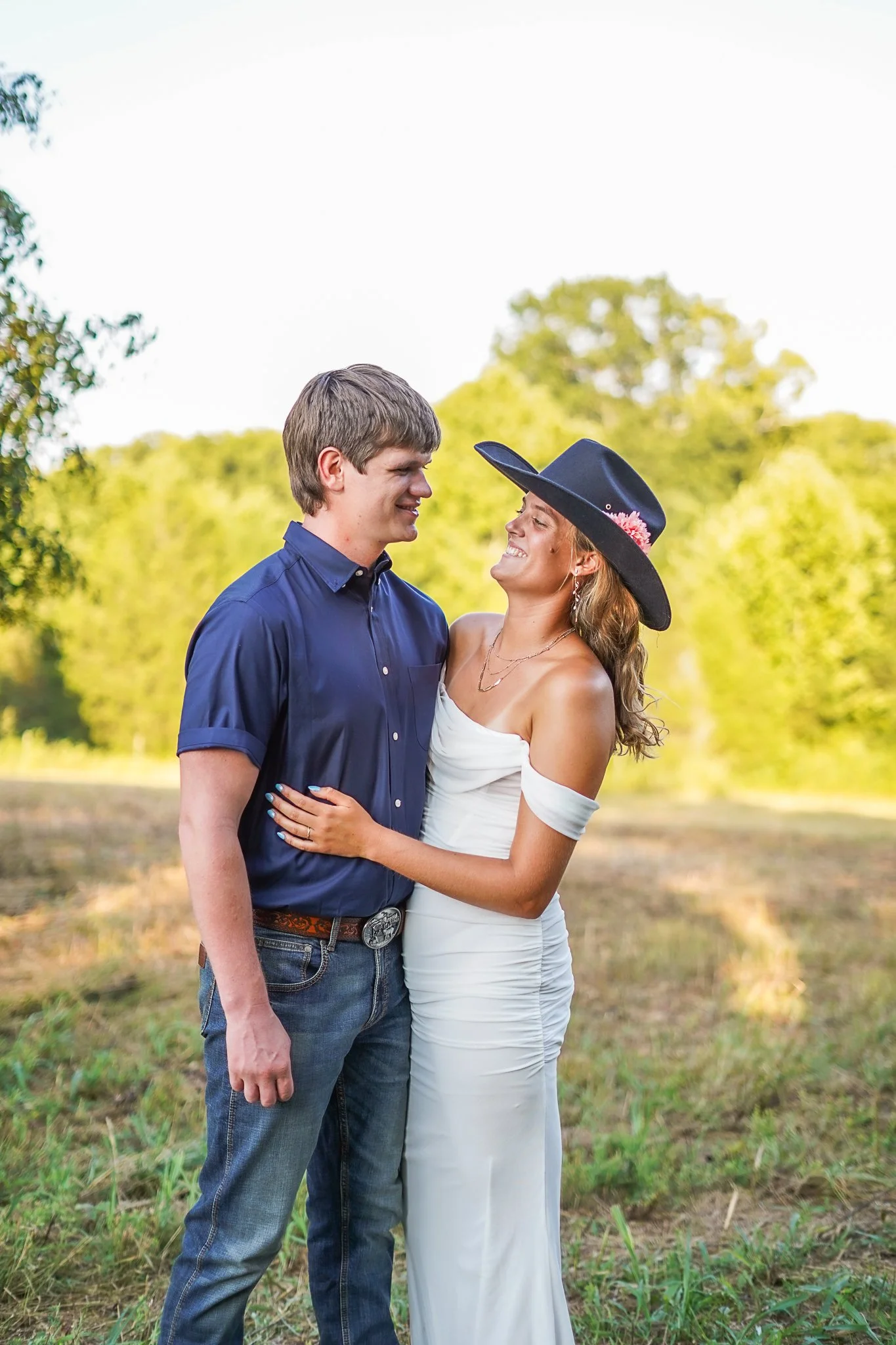 A man and woman are standing close together outdoors, smiling and looking at each other. The man is wearing a blue shirt and jeans, and the woman is wearing a white dress, a black wide-brimmed hat with a pink flower, and earrings. They are surrounded
