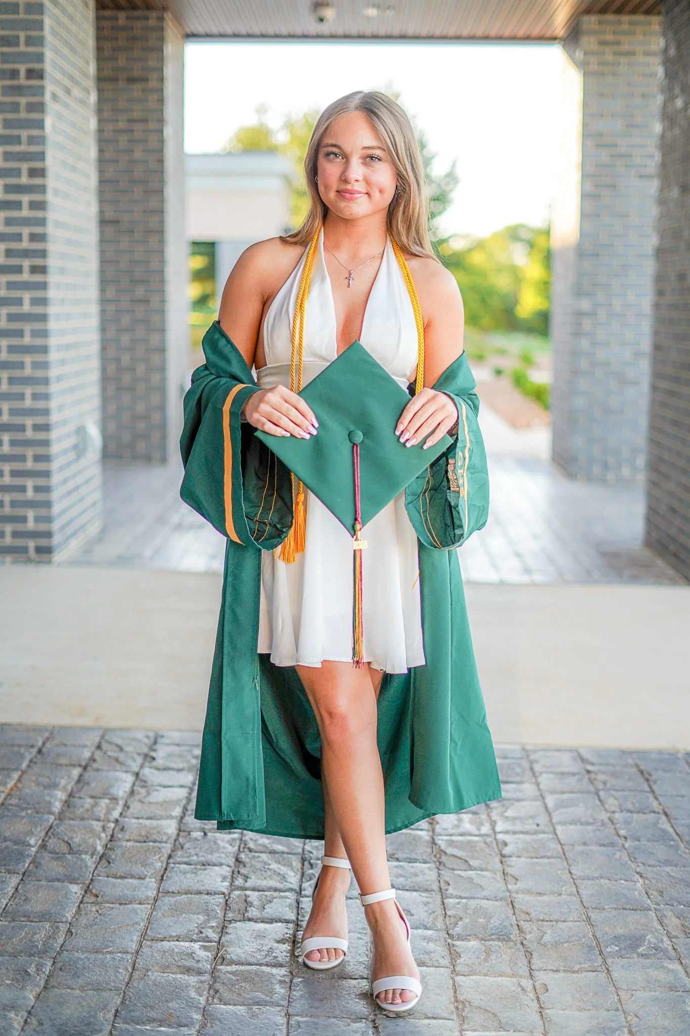 A young woman in a white graduation dress with a green gown and cap draped over her shoulders stands outside on a cobblestone path. She is smiling, holding her cap in her hands, and wearing white high heels. The background features a brick building a