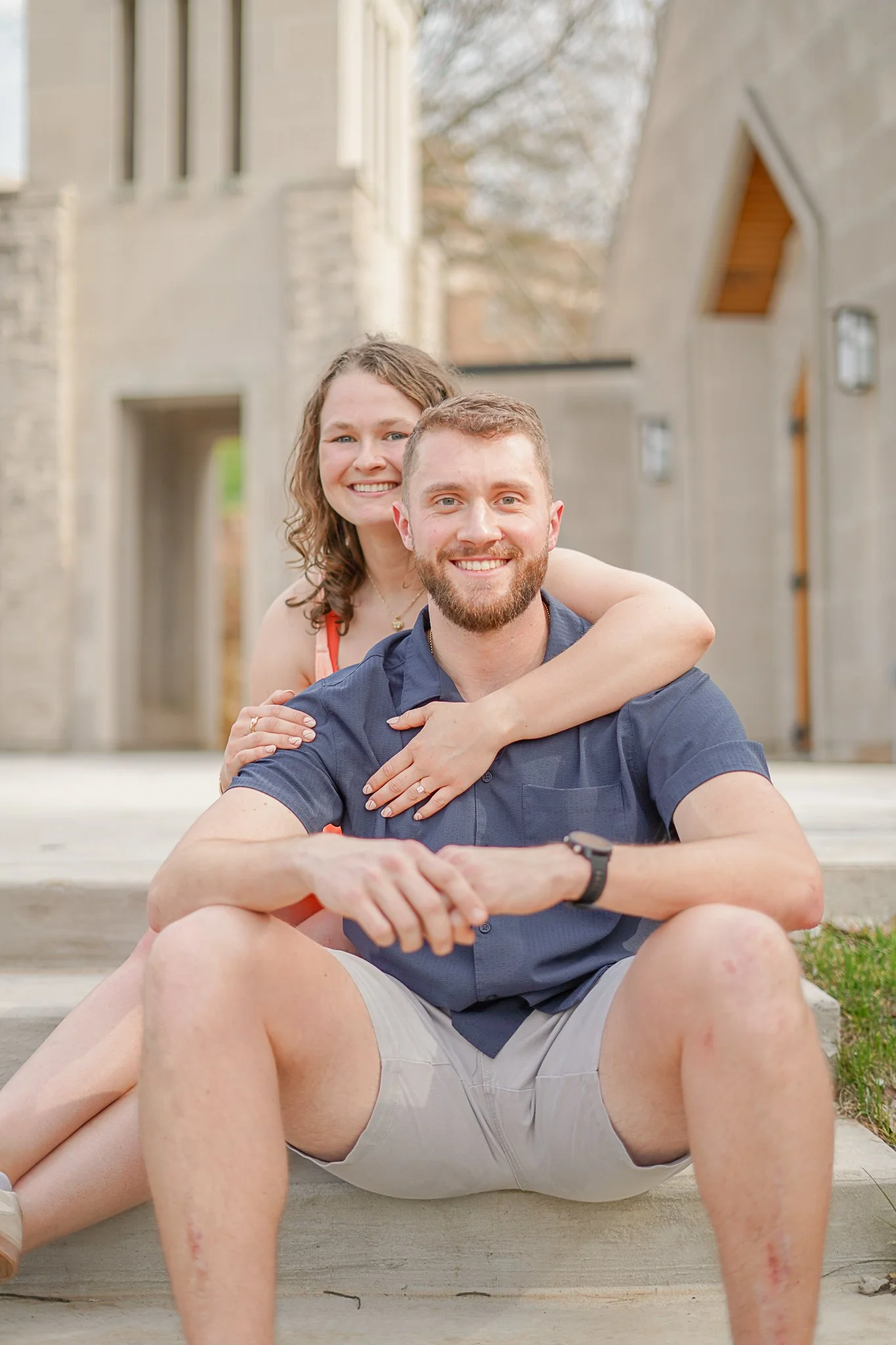 A smiling woman with curly hair showing a peace sign behind a smiling man with a beard, sitting on steps outside a house.