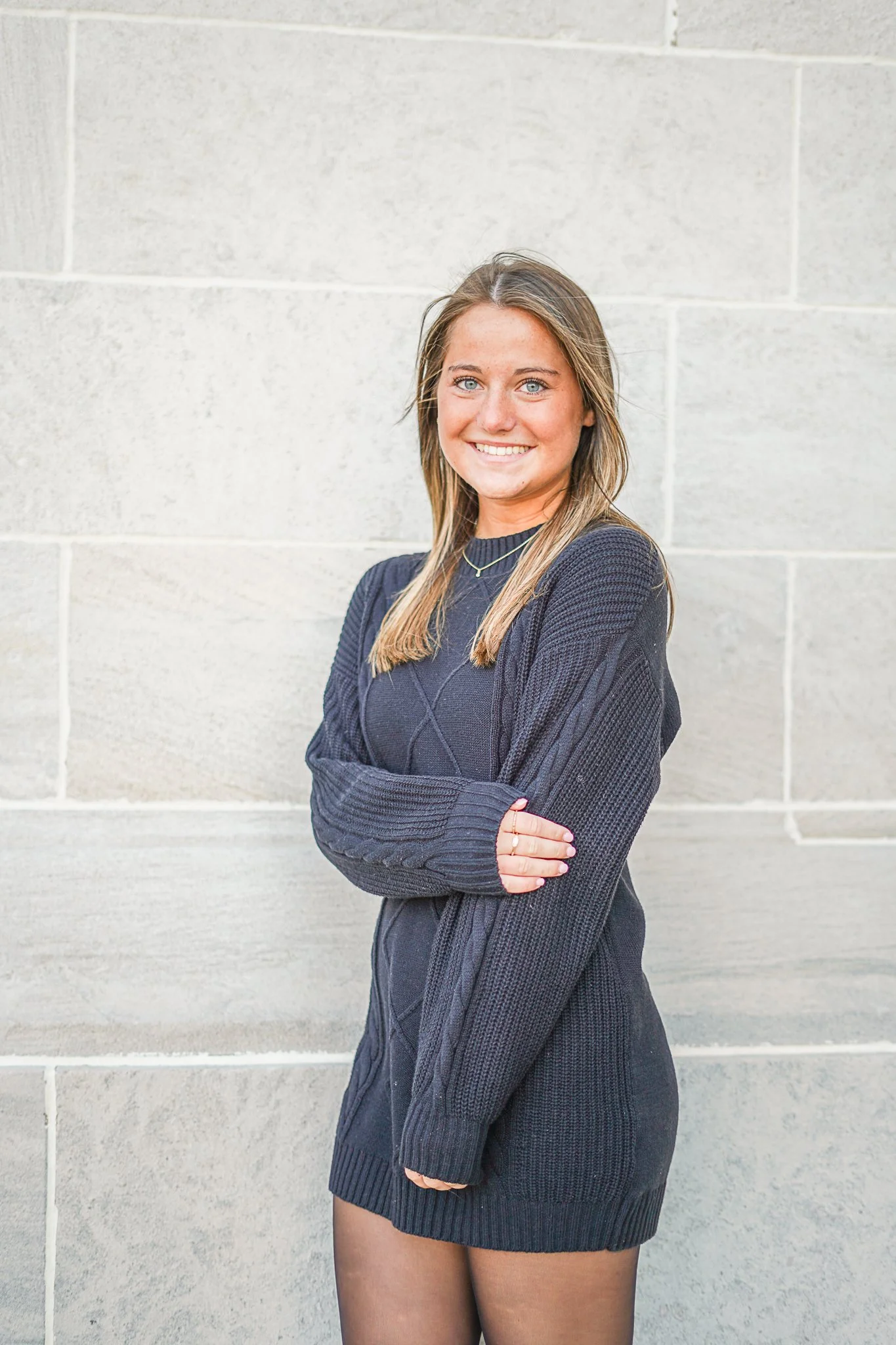 A woman smiling with crossed arms, wearing a black sweater, black skirt, and thigh-high stockings, standing against a light gray brick wall.