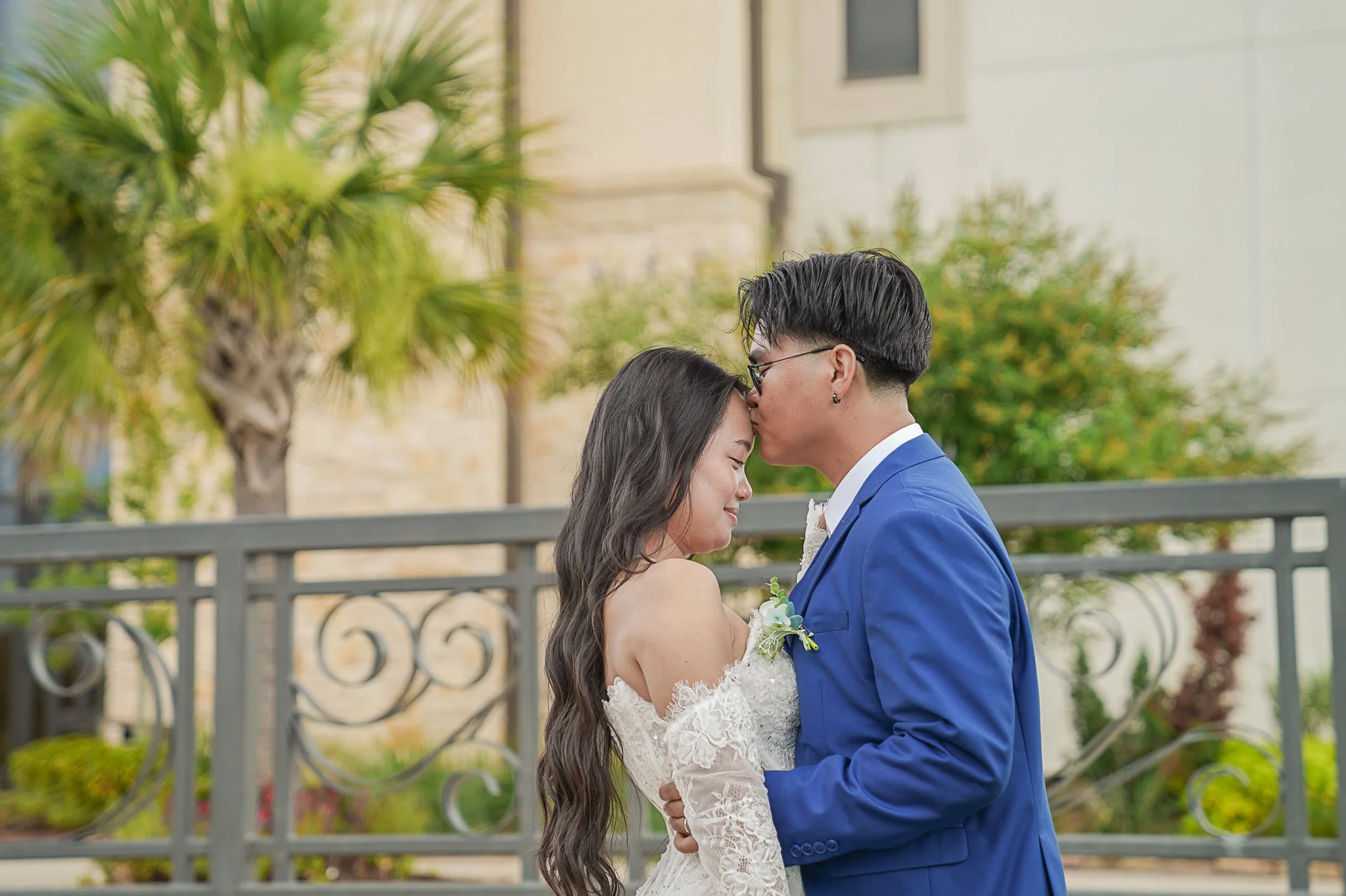 A couple dressed in wedding attire sharing a kiss outdoors with trees and a decorative metal fence in the background.