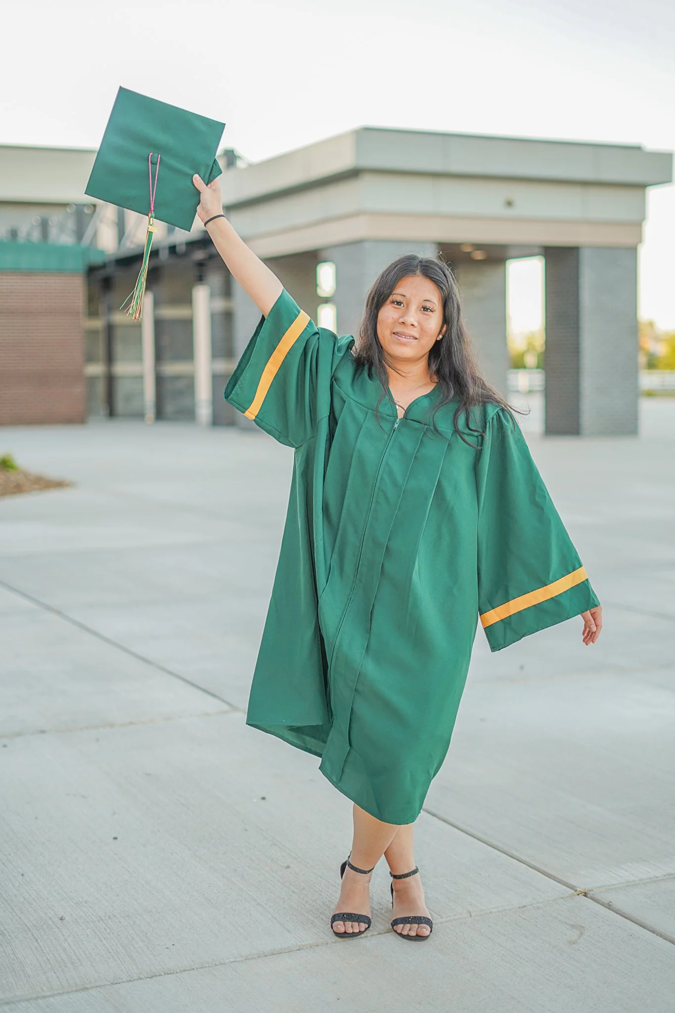 A young woman in a green graduation gown holds a graduation cap in the air outdoors near a modern building, celebrating her graduation.