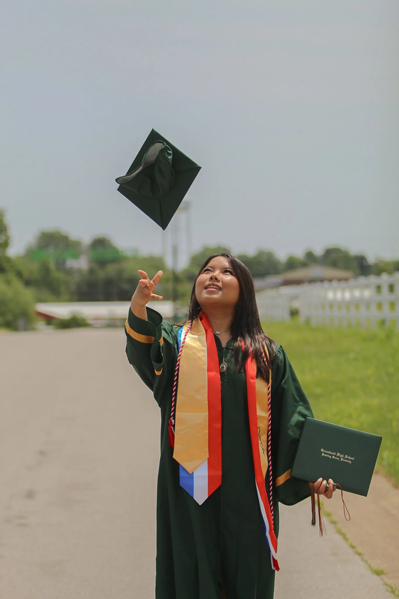 A young woman in a graduation gown and cap tossing her cap into the air outdoors on a cloudy day, holding a diploma and a green folder.