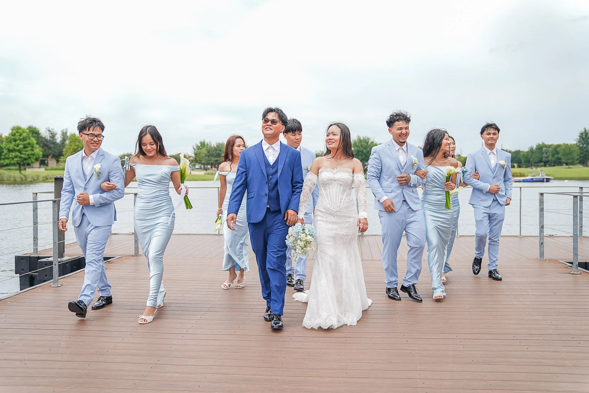 A wedding party walking on a wooden dock near a lake, with greenery and trees in the background under a cloudy sky.