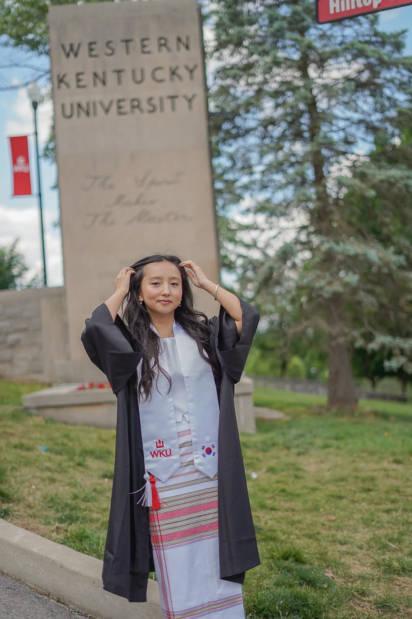 A young woman in graduation attire standing outdoors, adjusting her hair, with a university campus sign for Western Kentucky University and other signs and trees in the background.