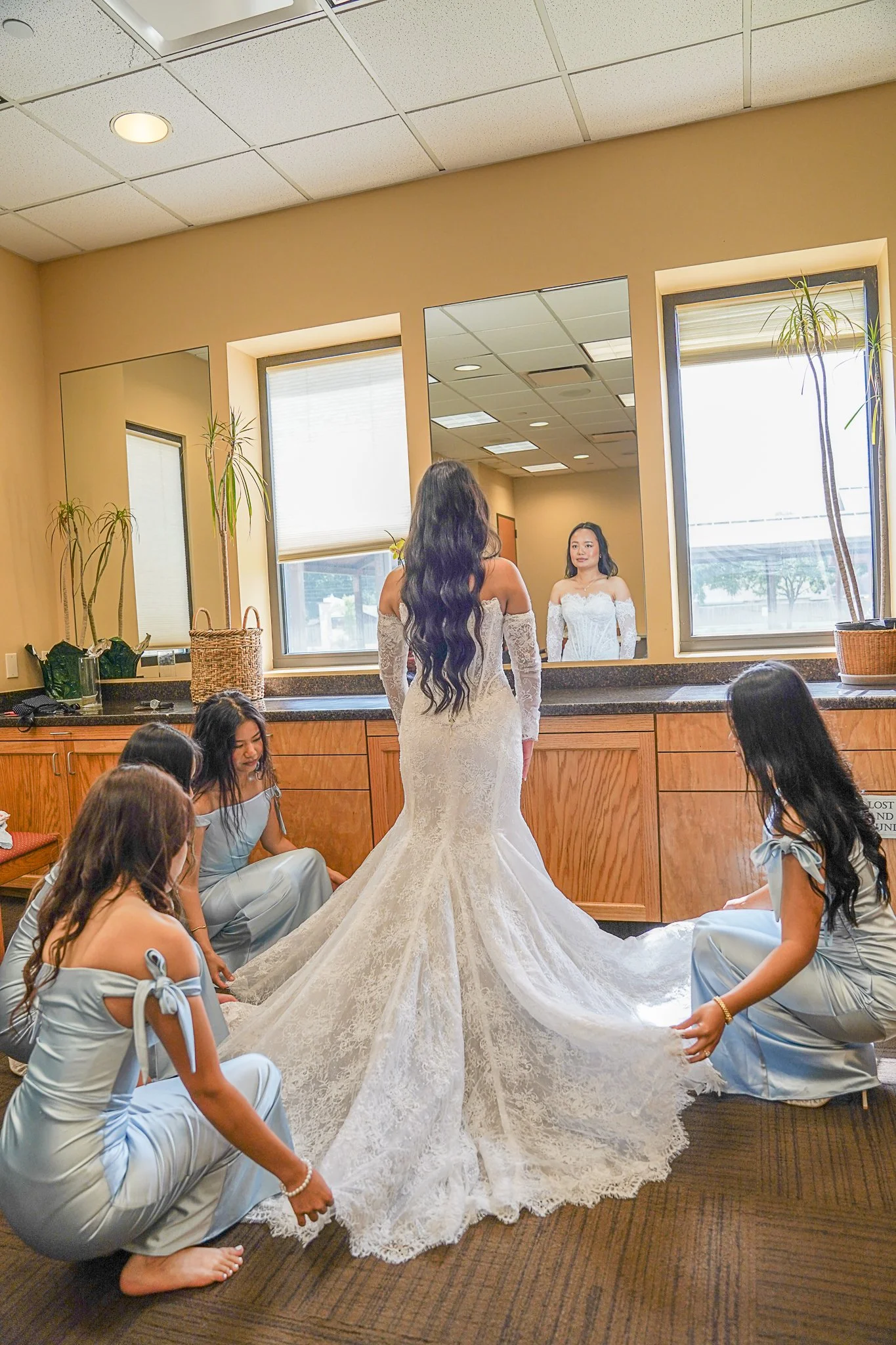 A bride in a white lace wedding gown with long train standing in front of a large mirror, surrounded by bridesmaids in light blue dresses sitting on the floor.