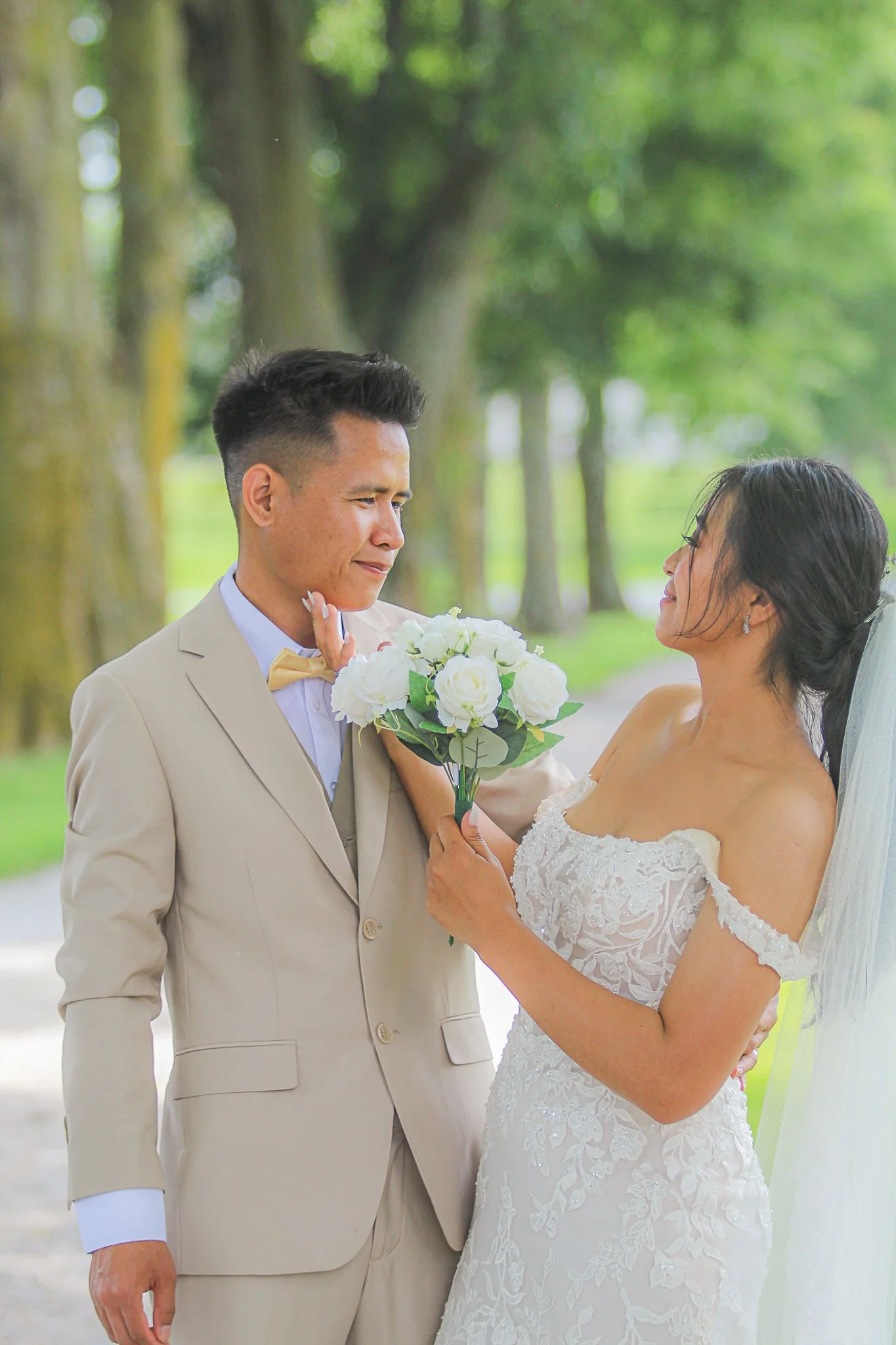 A wedding scene with a young man in a beige suit and a woman in a white lace wedding dress outdoors in a green park. The woman is handing the man a bouquet of white flowers.