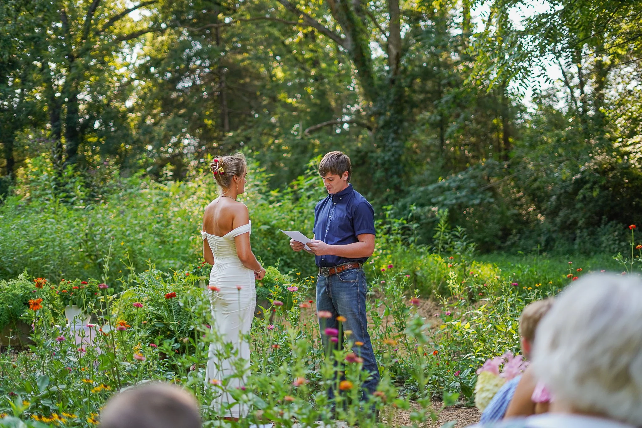 A woman in a white off-shoulder dress standing in a garden with colorful flowers, facing a young man in a blue shirt who is reading from a paper. Other people are partially seen in the foreground, surrounded by tall trees and greenery.