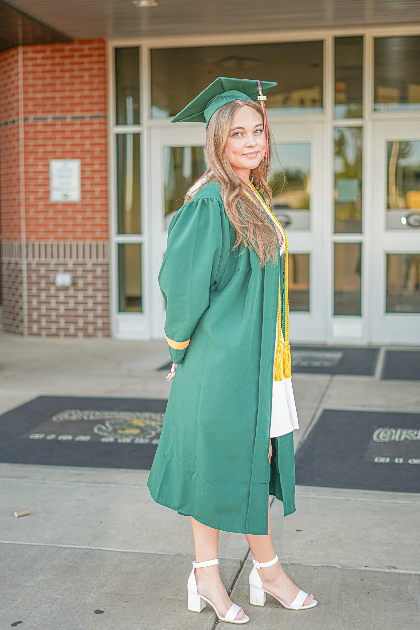 A young woman in a green graduation gown and cap with pink and yellow cords, standing outside a building with glass doors and brick wall, wearing white high-heeled shoes.