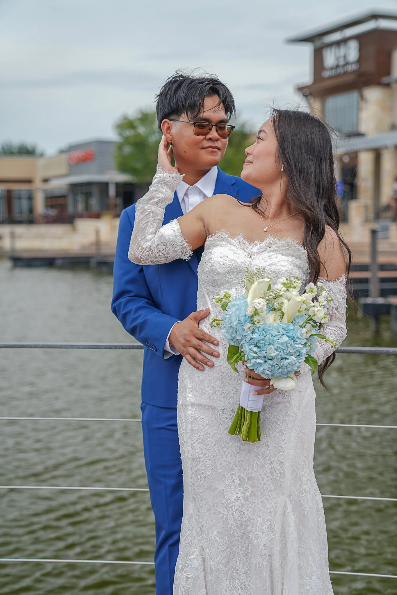 A bride in a white lace wedding dress and a groom in a blue suit, holding a bouquet of blue and white flowers, standing on a boat dock with water in the background.